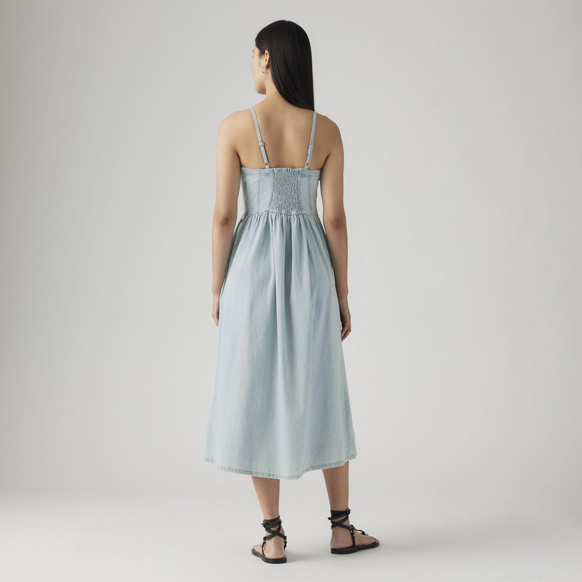 A woman with long dark hair, facing away, wears the Levis Alessie Bustier Dress—a light blue, sleeveless, midi A-line dress—with black strappy sandals against a plain light background.