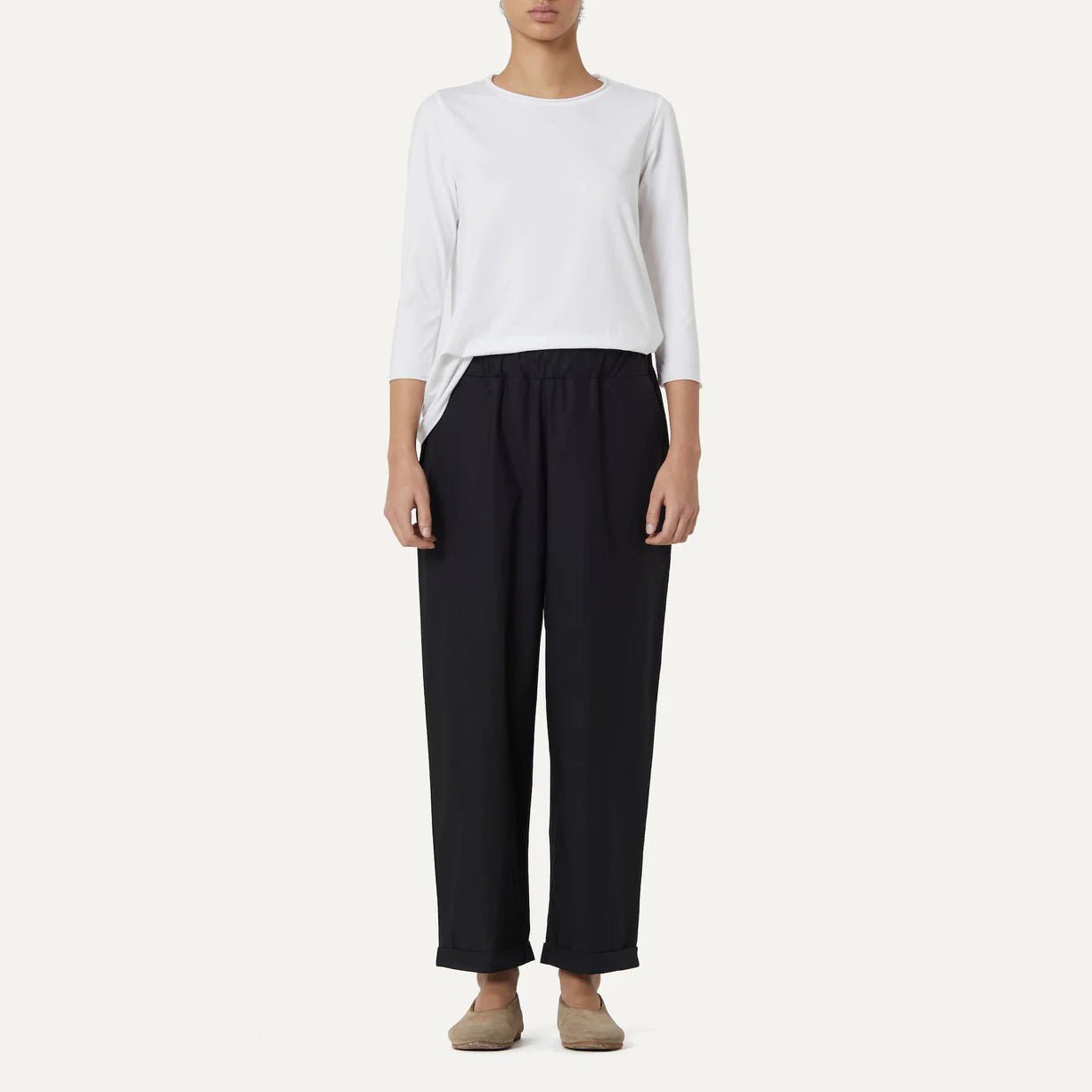 A person dressed in a white cotton poplin long-sleeve shirt, Labo Art's Panta Vela Pant, Clara in black with an elastic waistband, and beige flat shoes stands against a plain light background.