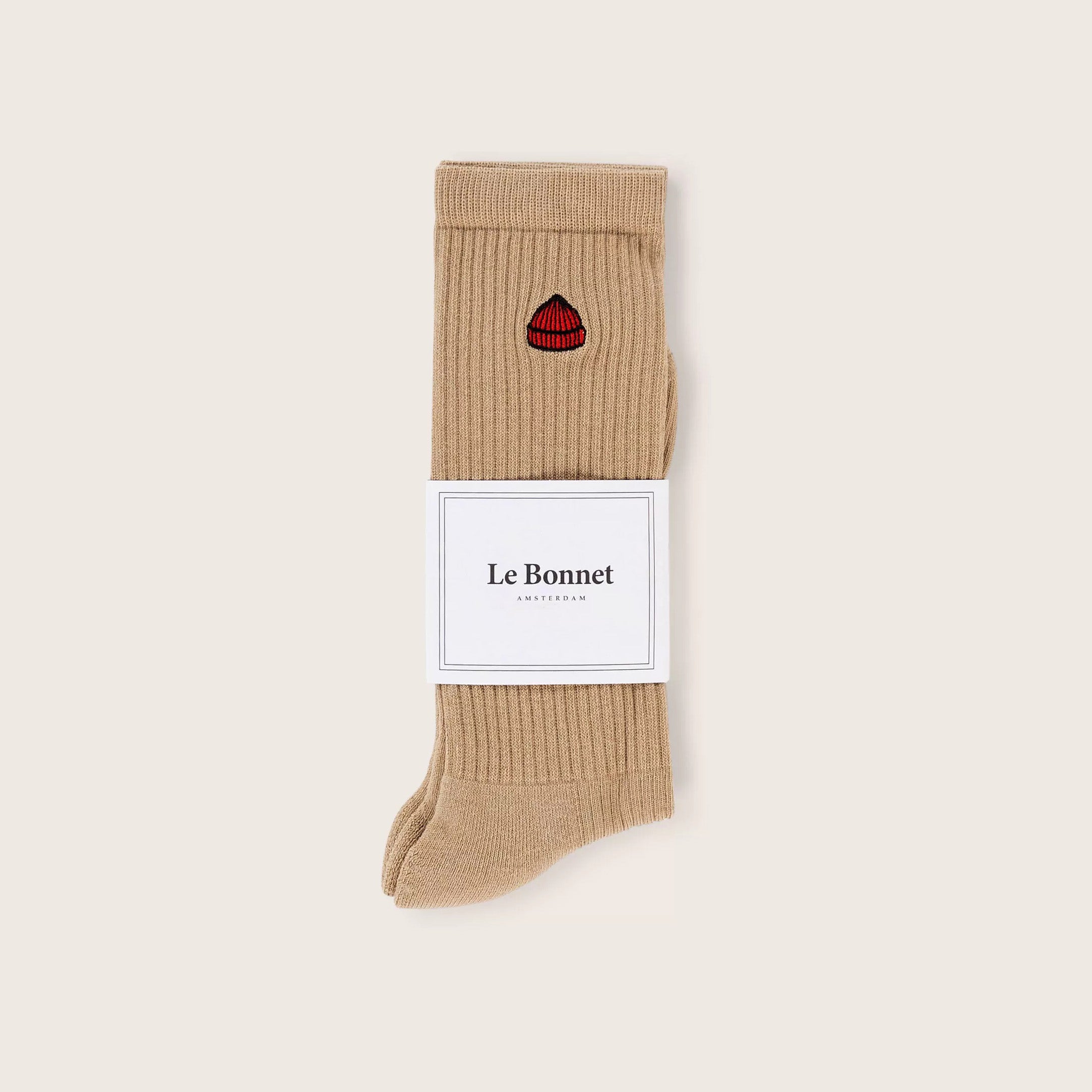 A pair of Le Bonnet Tube Socks in Sand, crafted from a soft cotton blend with ribbed texture, features a small red lips patch and is wrapped with a white Le Bonnet label.