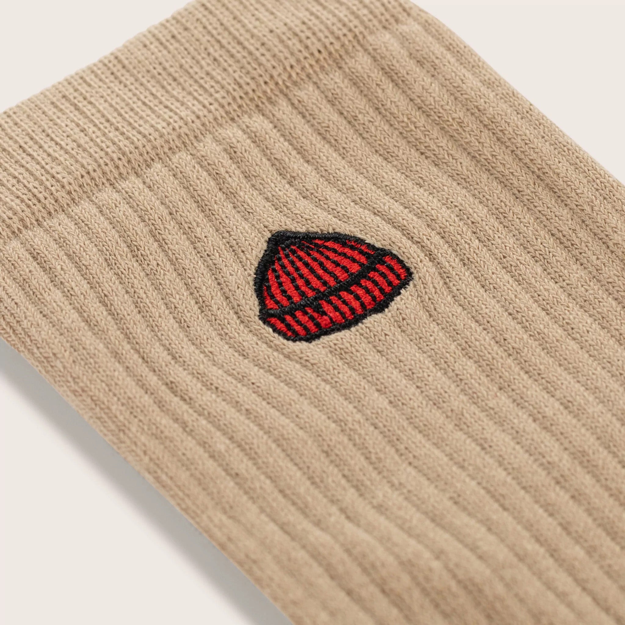 Close-up of Le Bonnet's Tube Sock in Sand, a beige ribbed knit crafted from a soft cotton blend and featuring an embroidered red and black acorn motif.