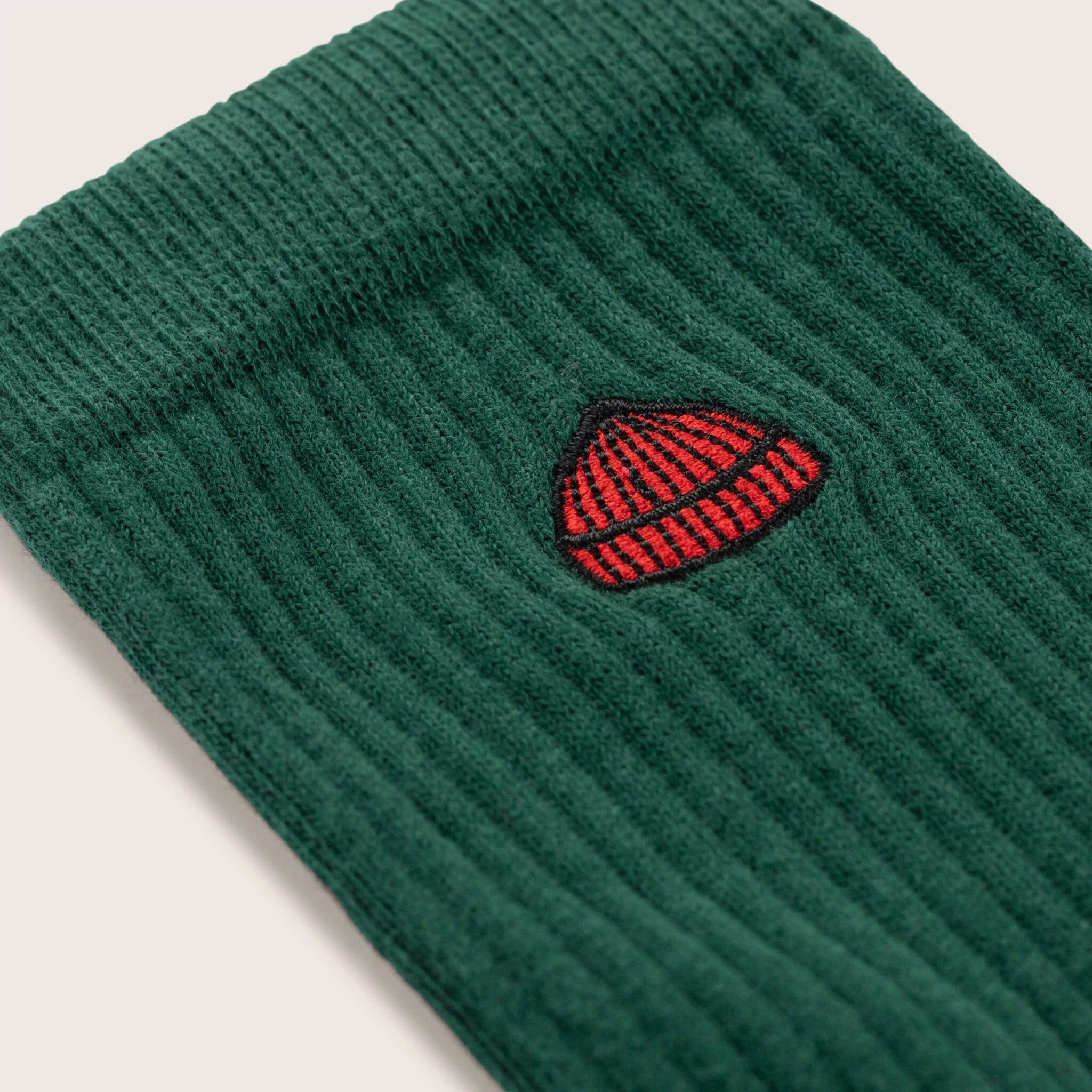 Close-up of Le Bonnet's Tube Sock in Pine, made from a soft cotton blend with green ribbed fabric, finished with a small red beanie embroidery.
