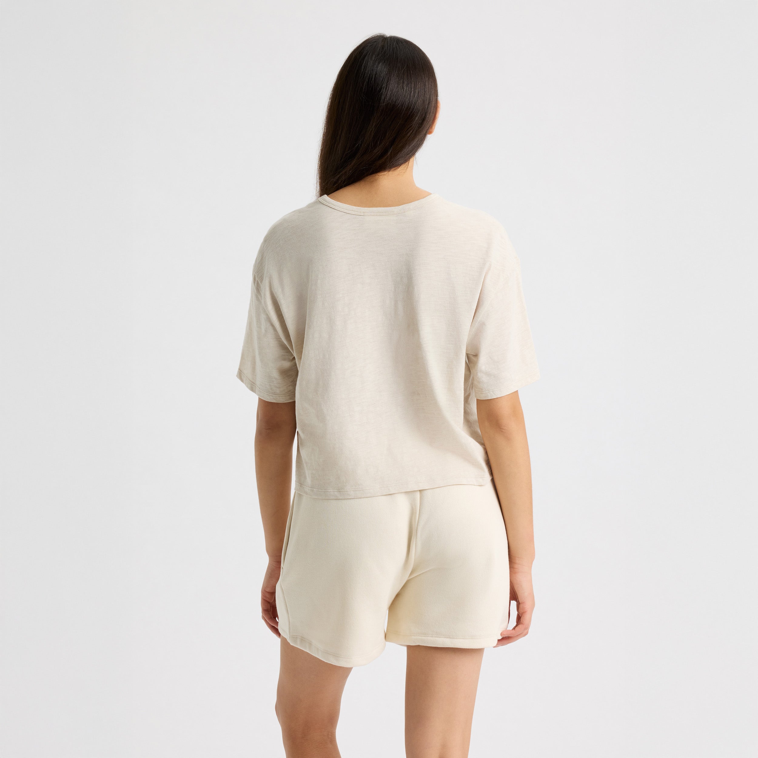 A person with long dark hair is wearing Rohnisch's beige Slub Cropped Tee and shorts, standing with their back to the camera against a plain white backdrop. This T-shirt offers a boxy fit ideal for relaxed, laid-back days.
