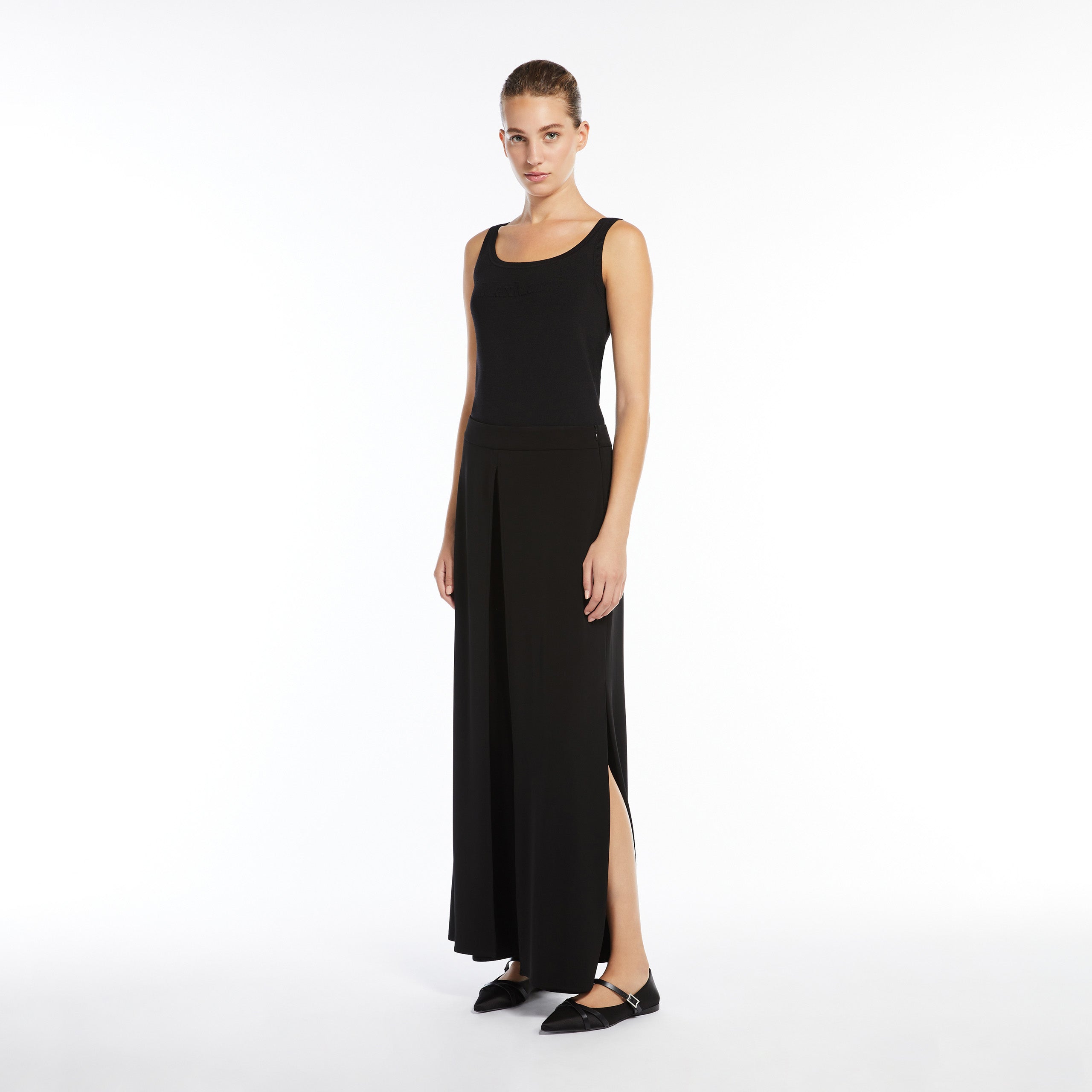 A woman stands against a white background wearing the Max Mara Pentola Sweater Tank Top, a long black viscose crêpe skirt with side slits, and black flat shoes.