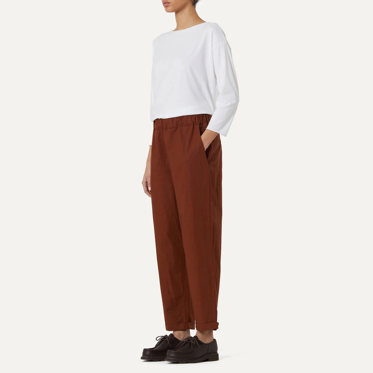 A person wearing a white long-sleeve shirt, Labo Art's "Panta Vela Pant, Clara," styled with an elastic waistband and in brown, stands in profile against a plain background, paired with black shoes.