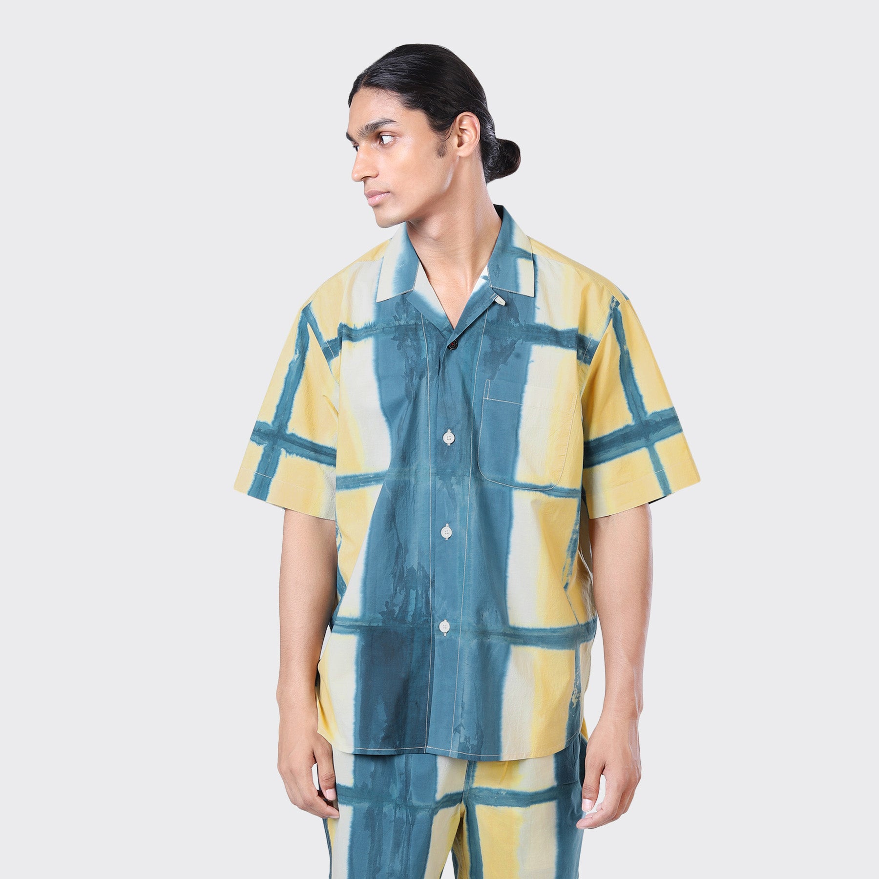A person wears the Kardo Ronen SS Shirt in Yellow Clamp Dye, featuring a yellow and teal plaid pattern, with matching pants, looking to the side against a plain background.