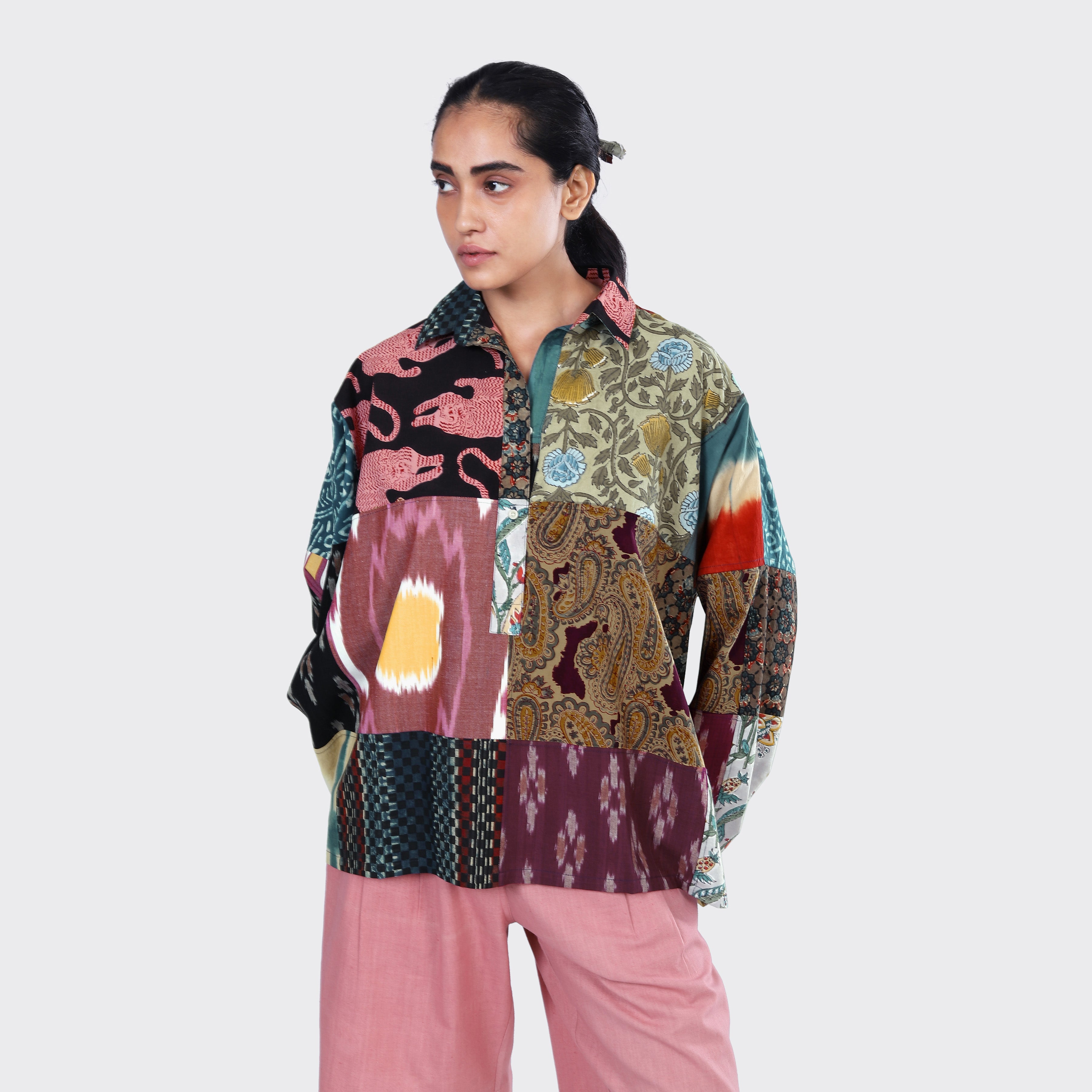 A person wears Kardo's Fanette Overshirt, Patchwork 07—a loose, multicolored patchwork shirt—paired with pink pants, standing against a plain light background.
