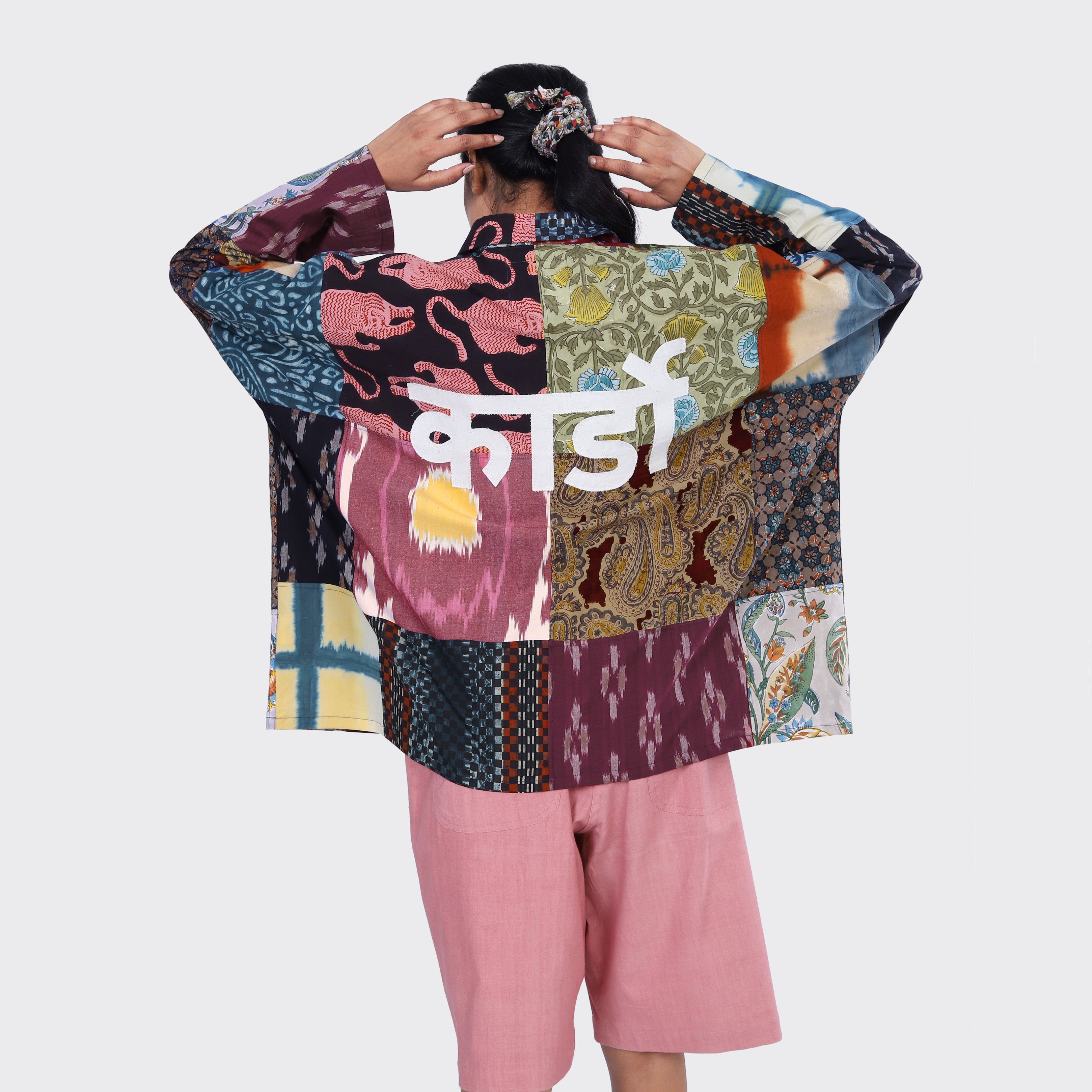 Wearing the Kardo Fanette Overshirt, Patchwork 07 with Hindi text on the back and pink shorts, a person faces away adjusting their hair—an eye-catching SS25 look.