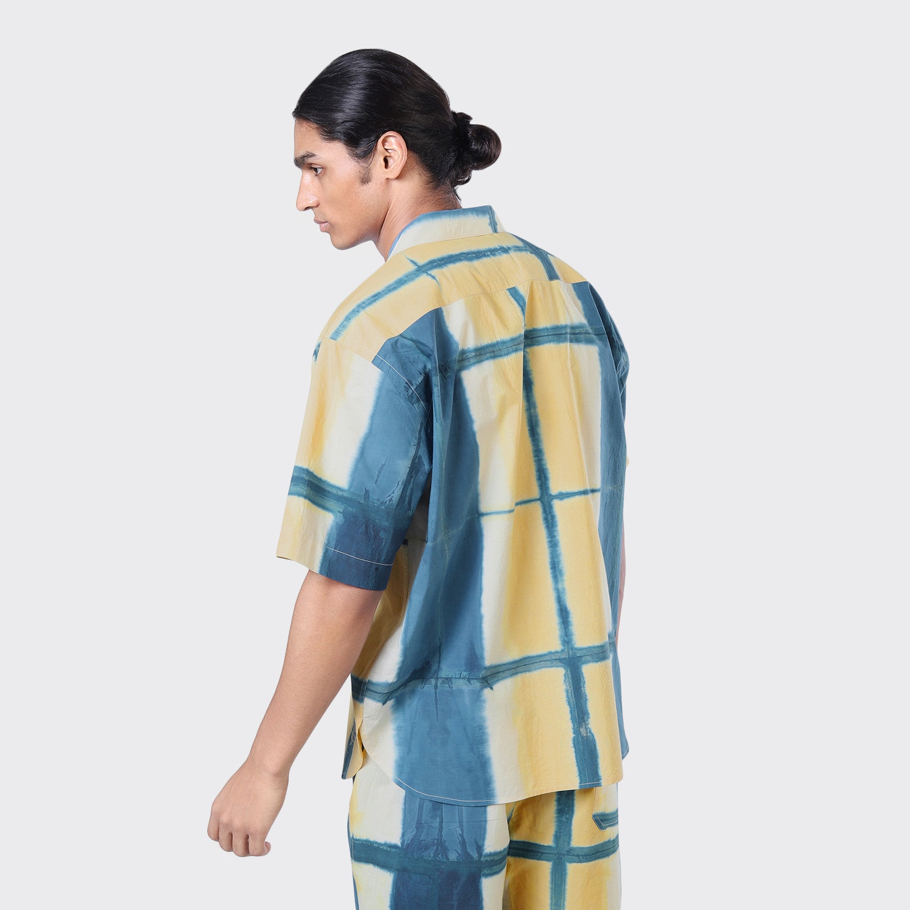 A man with dark hair in a low bun is seen from the back against a plain light background, wearing the Kardo Ronen SS Shirt in Yellow Clamp Dye with matching pants.