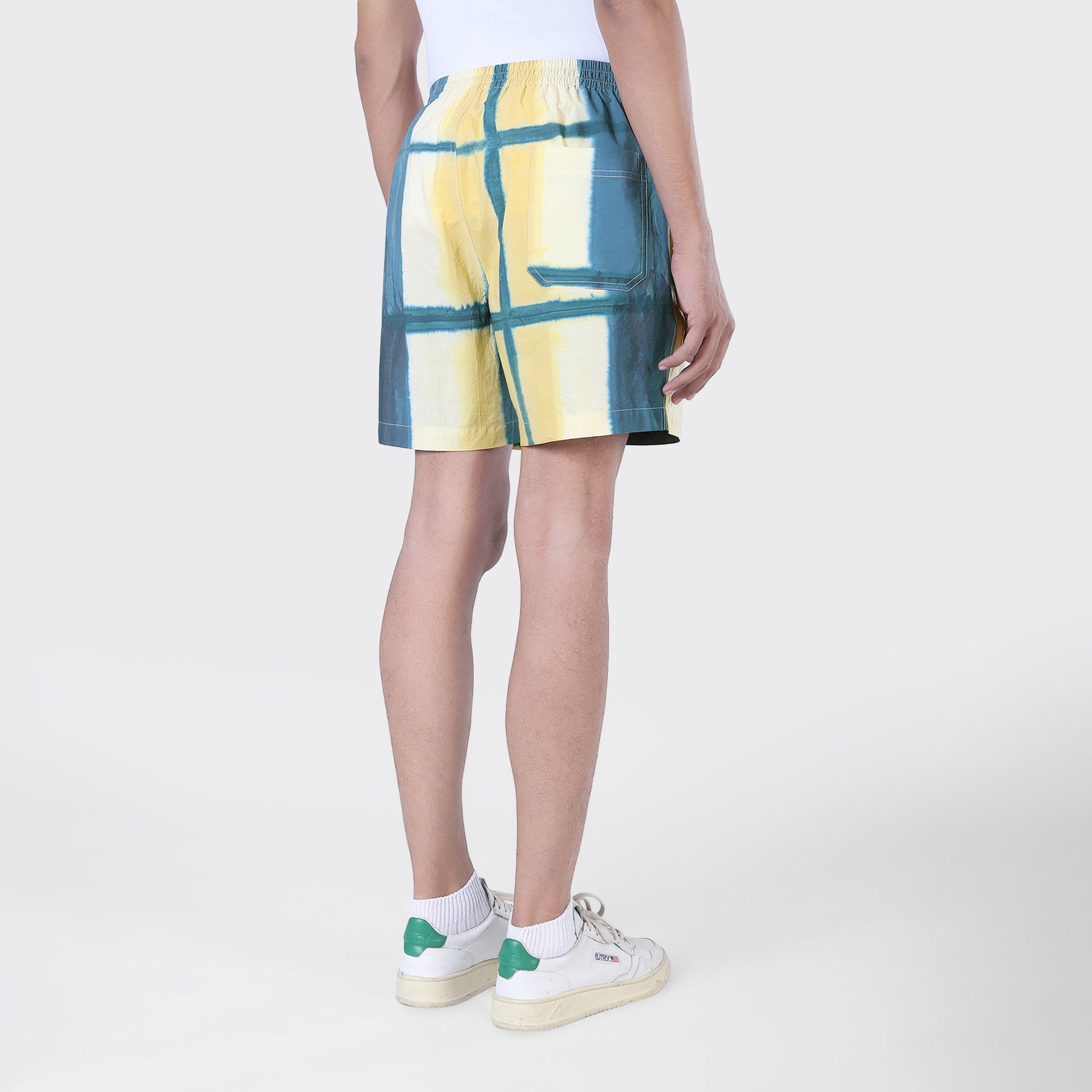 A person stands on a white background wearing a white tank top, Kardo Olbia Shorts in Yellow Clamp Dye, white socks, and white sneakers with green accents.