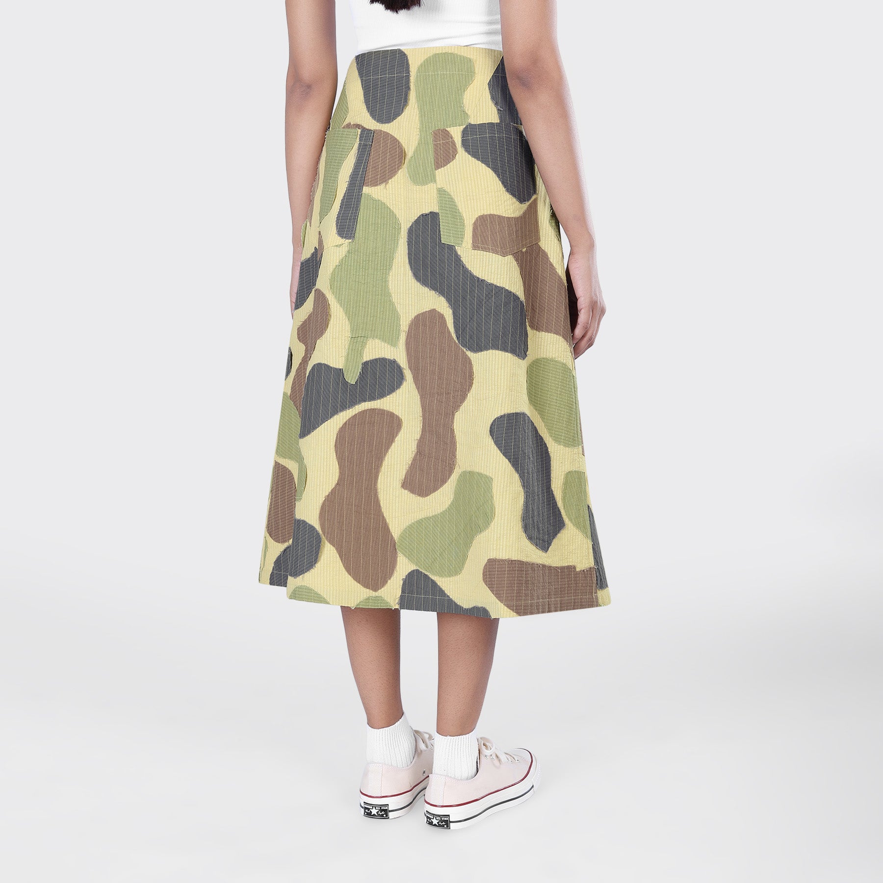Shown from the back against a plain background, a person wears a white top, Kardo’s Yuki Skirt in Patchwork Camo, plus white socks and sneakers.
