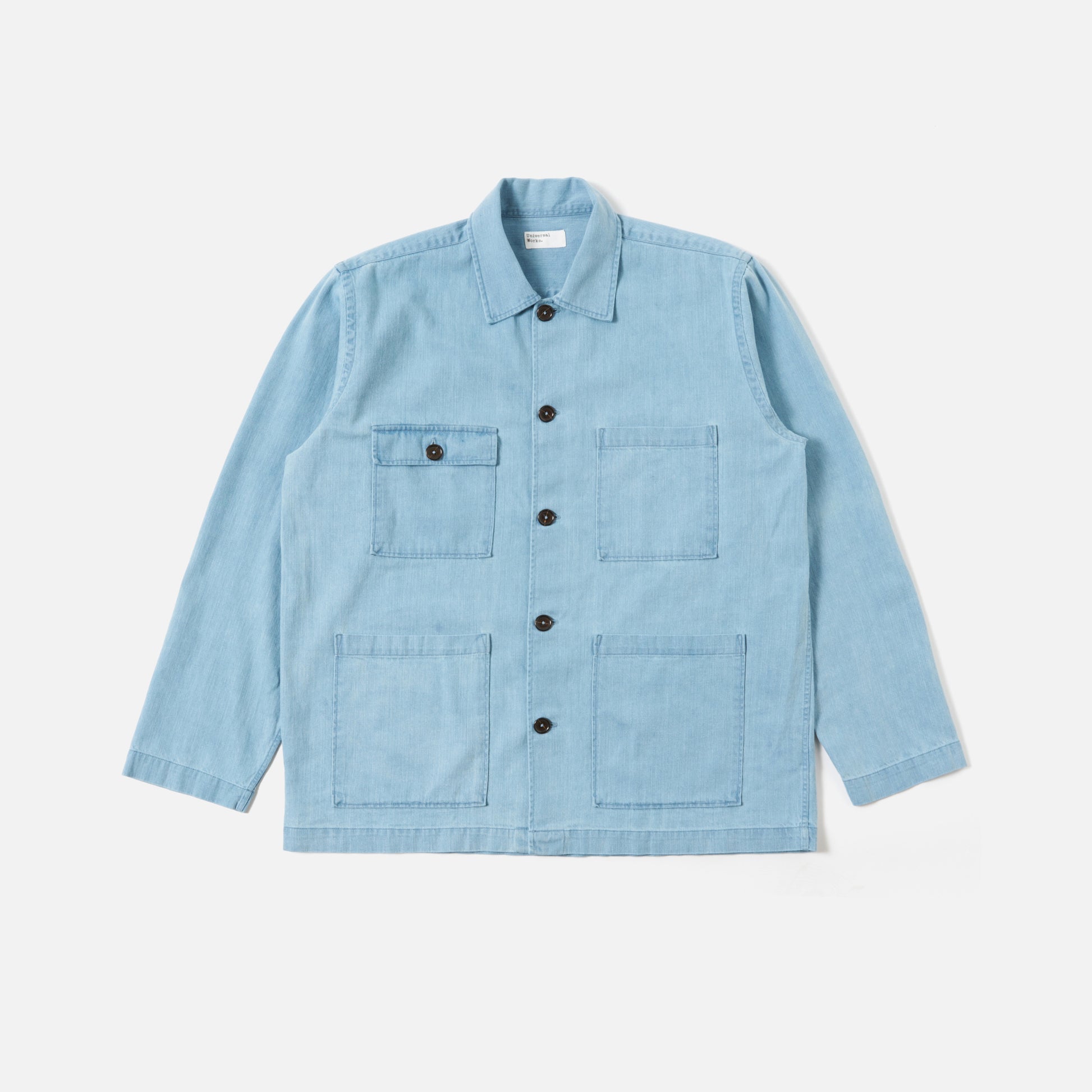 The Universal Works 8 Oz Organic Denim Dockside Jacket in Washed Indigo features long sleeves, four front pockets, and black buttons. Pictured laid flat on a plain white background.