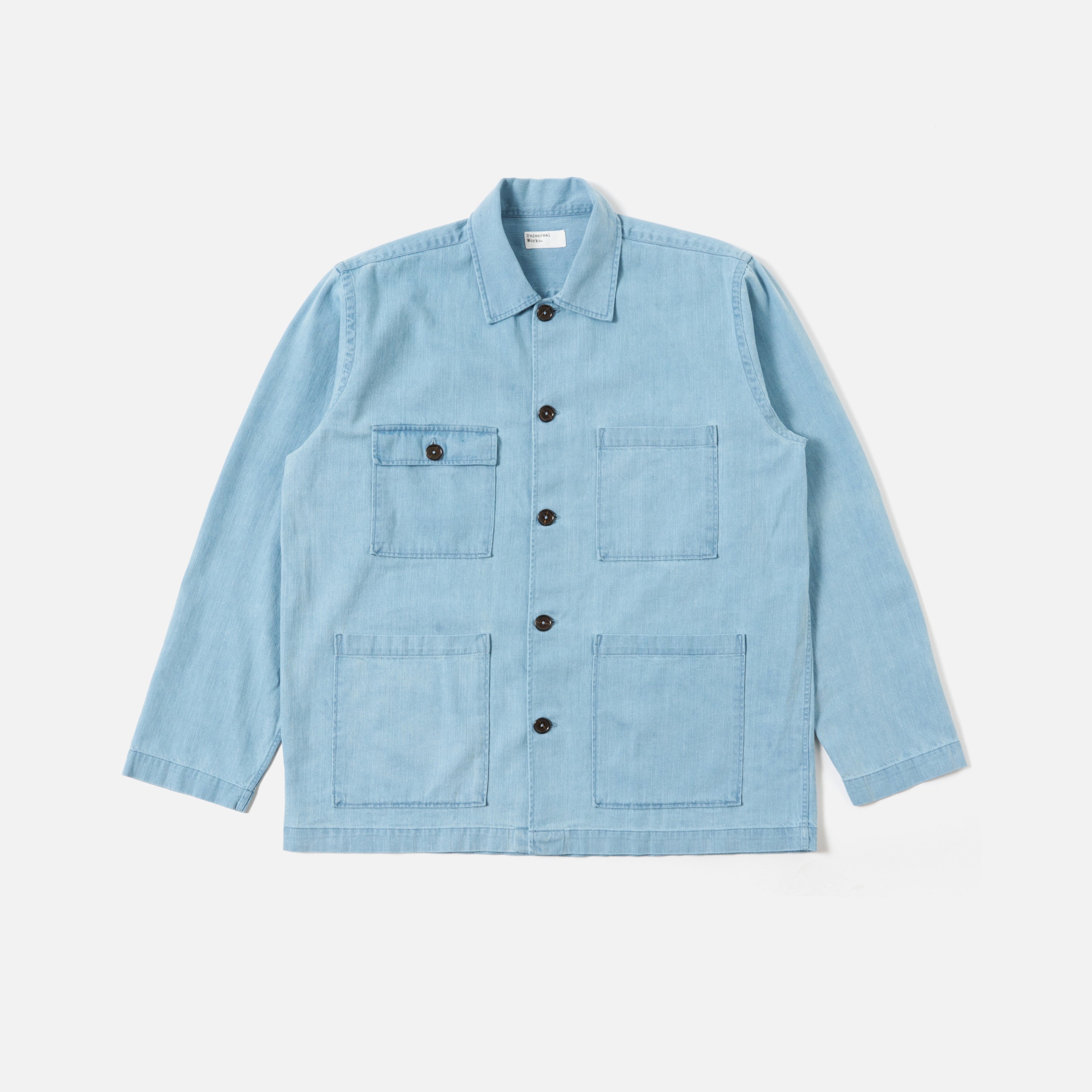 The Universal Works 8 Oz Organic Denim Dockside Jacket in Washed Indigo features long sleeves, four front pockets, and black buttons. Pictured laid flat on a plain white background.