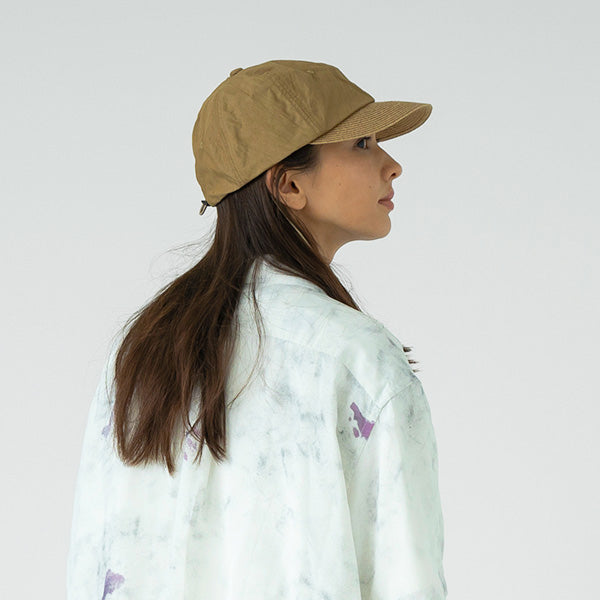 A woman wearing the Charidoor Cap by Sublime Roe and a light tie-dye shirt stands sideways, gazing right against a plain background.
