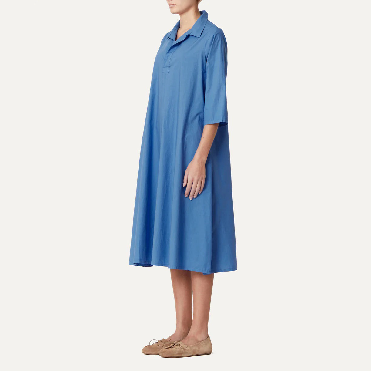 A person stands sideways against a plain background wearing the knee-length Abito Pepe Dress, Clara by Labo Art, featuring elbow sleeves in blue Poplin fabric, paired with brown shoes.