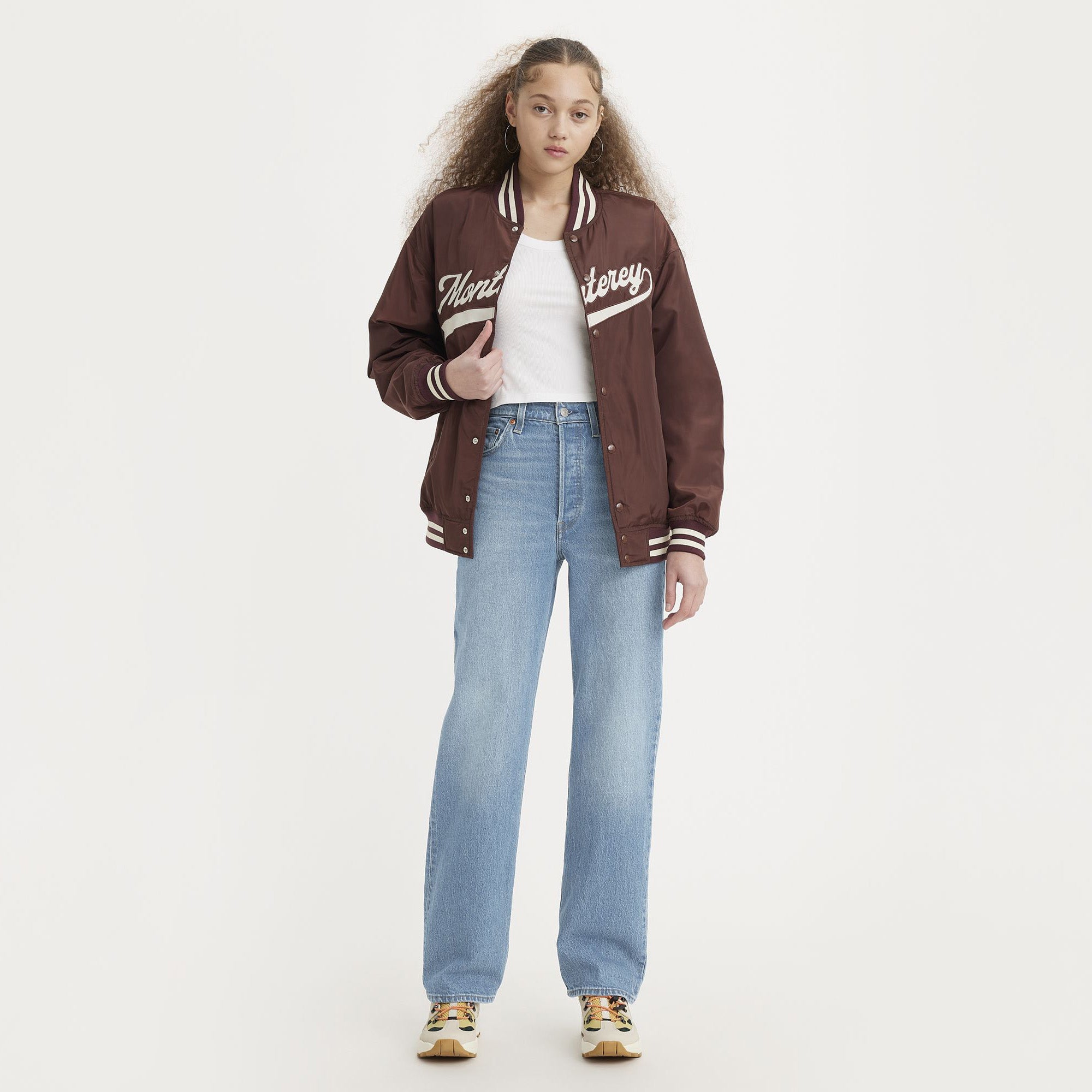 A young woman faces forward against a white background, wearing a maroon varsity jacket, white t-shirt, yellow sneakers, and Levis Ribcage Full Length light blue high rise jeans with a fitted seat and straight leg.