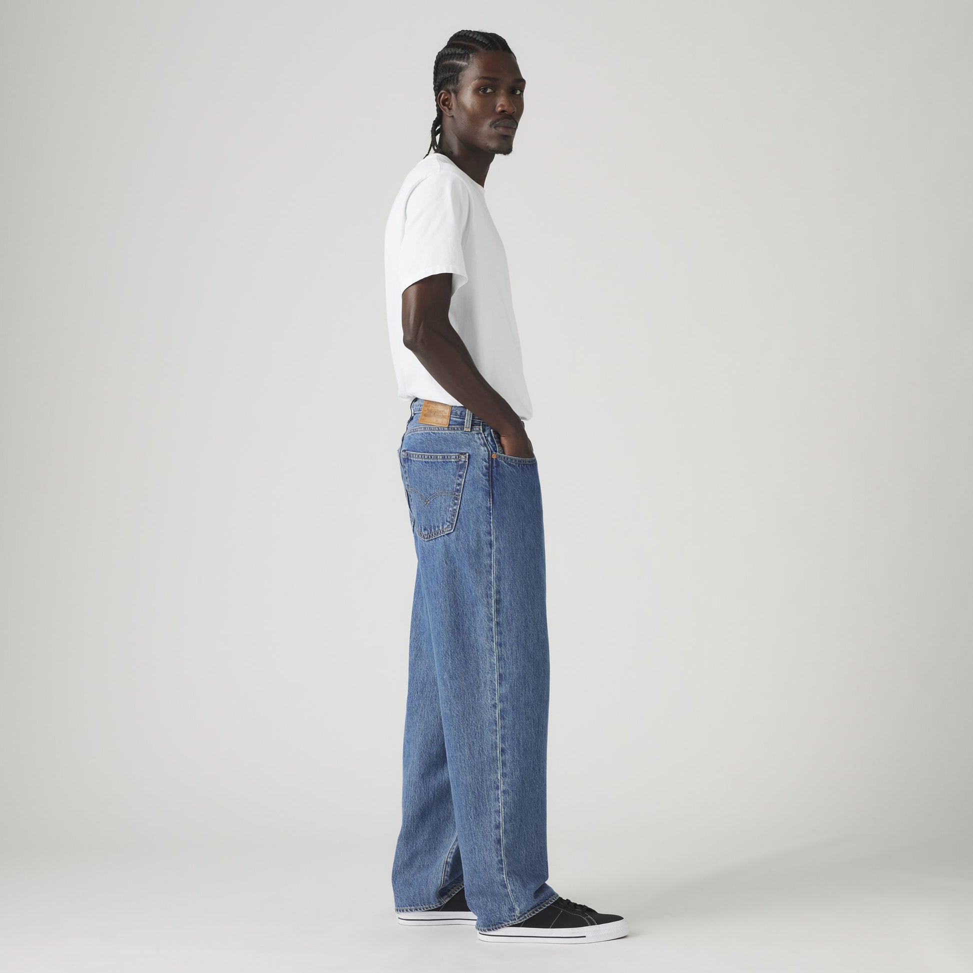 A person stands sideways against a plain light background wearing a white t-shirt, black sneakers, and Levis 578 Baggy non-stretch loose blue jeans.