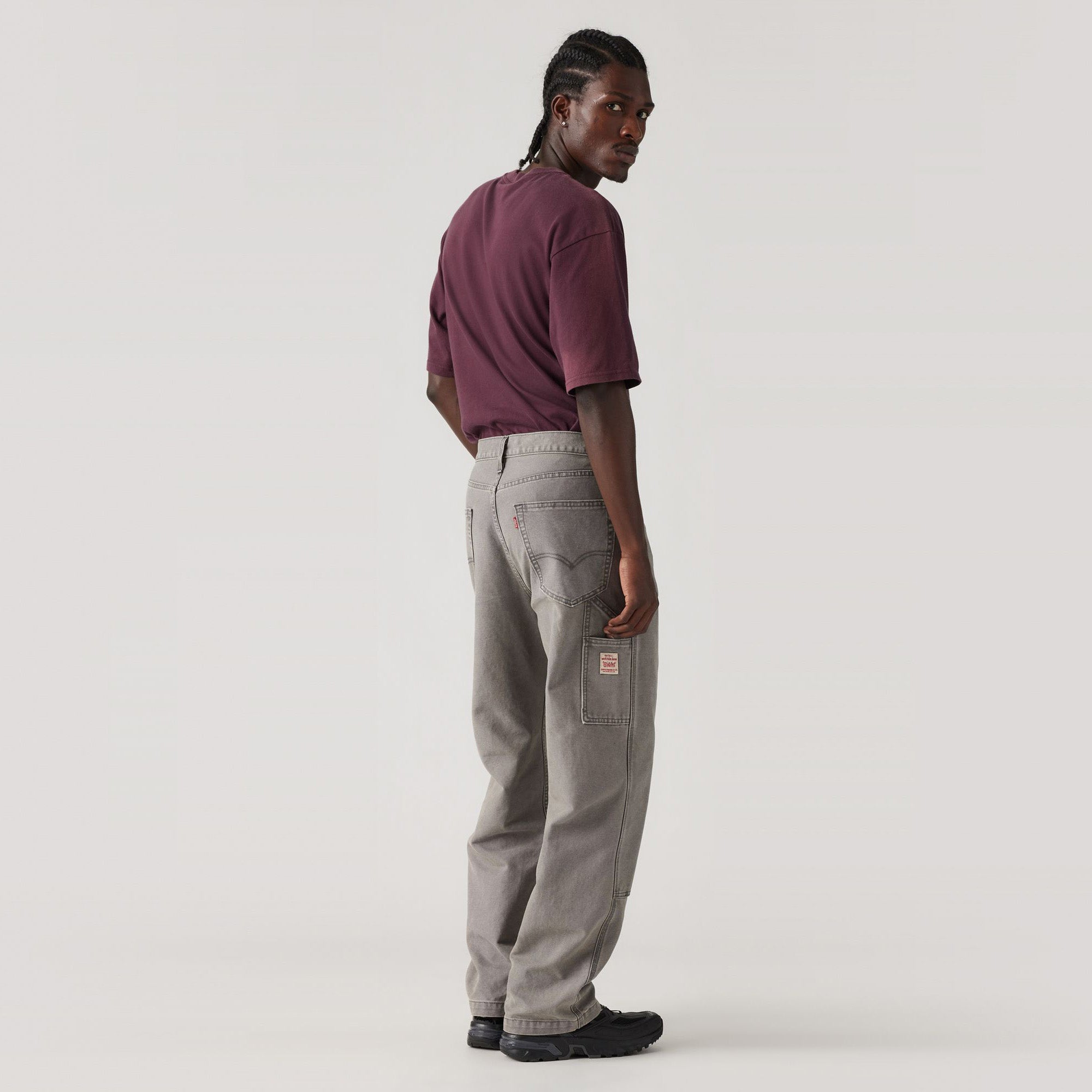A person stands sideways, wearing a maroon t-shirt, black shoes, and Levis 568 Loose Straight Double Knee gray cargo pants with visible stitching and pockets against a plain light background.