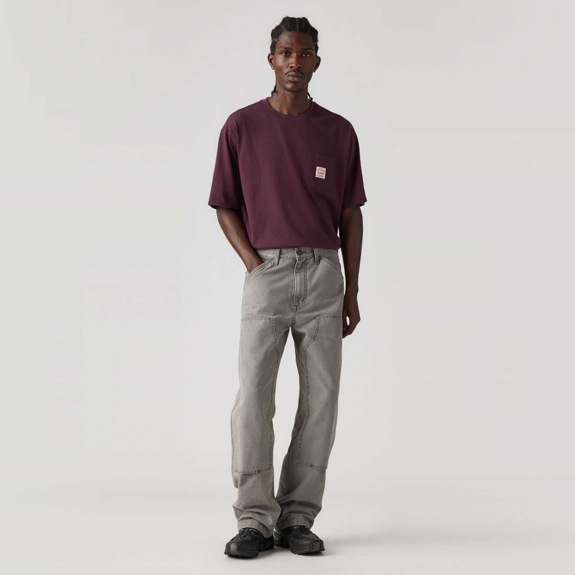 A man stands facing forward against a plain light background, wearing a maroon T-shirt, Levis 568 Loose Straight Double Knee light gray work pants, and black shoes.