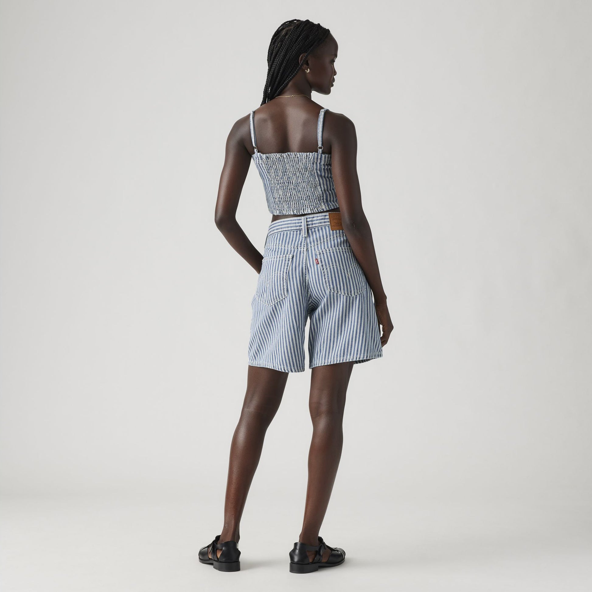 A person stands facing away, wearing a blue and white striped sleeveless top, Levis High Baggy Short, and black sandals against a plain light background.