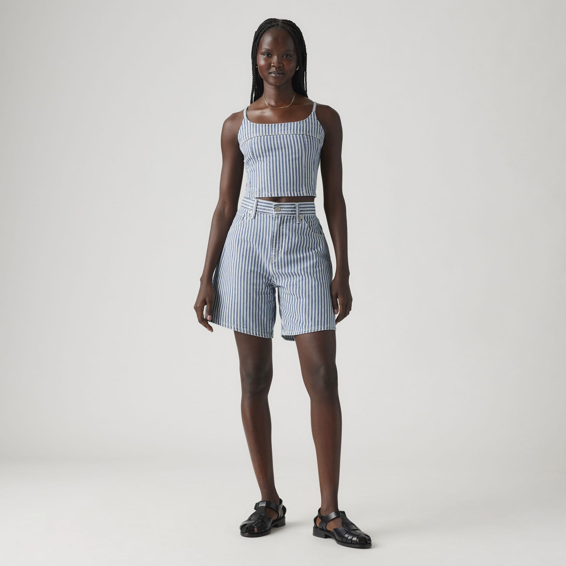 A woman stands against a plain background wearing a blue and white striped sleeveless crop top, Levi’s High Baggy Short with a belt, and black sandals.