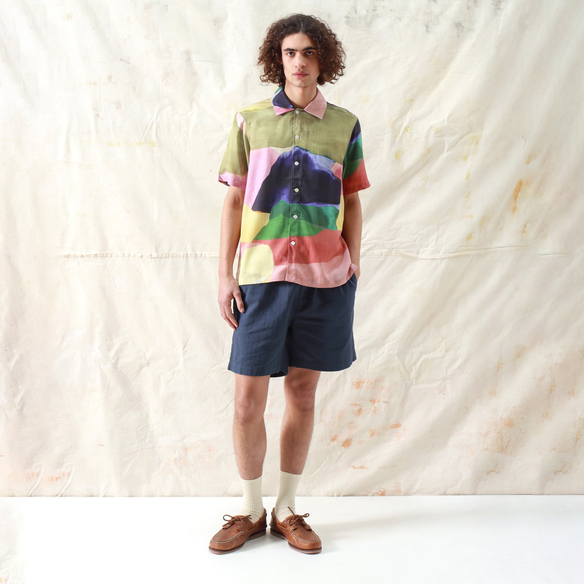 A person stands against a light, textured backdrop wearing the Far Afield Busey SS Shirt in Painted Landscape, paired with dark shorts, socks, and brown boat shoes.