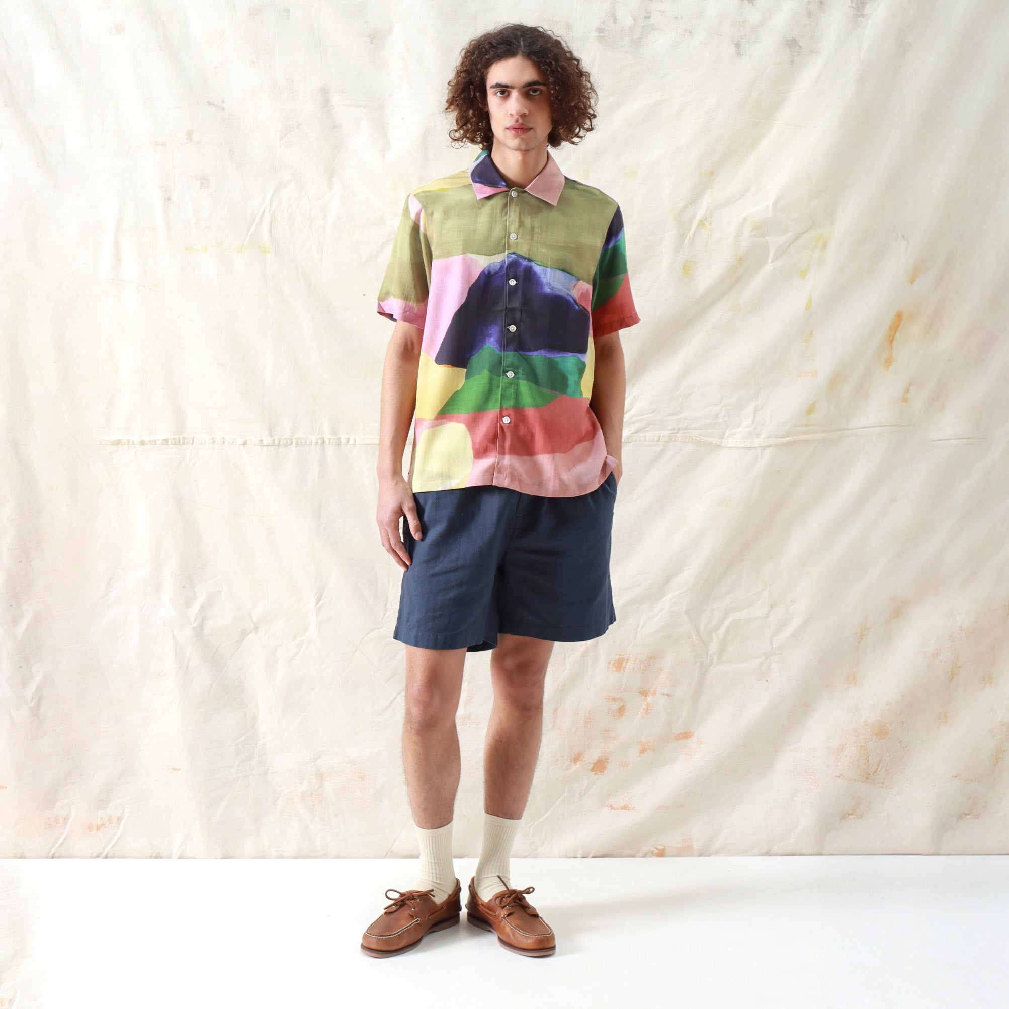 A person stands against a light, textured backdrop wearing the Far Afield Busey SS Shirt in Painted Landscape, paired with dark shorts, socks, and brown boat shoes.