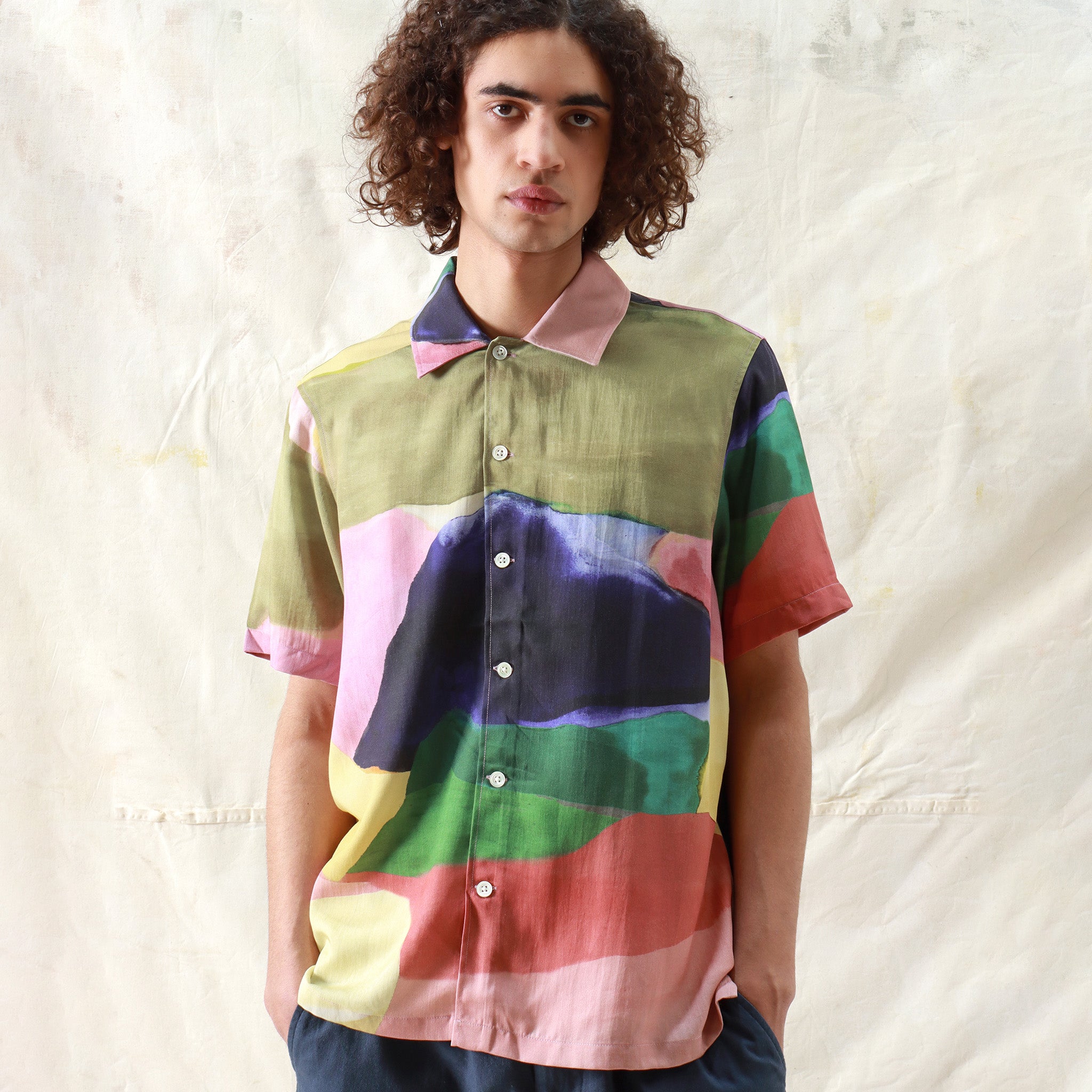 A person with curly hair wears the Far Afield Busey SS Shirt, Painted Landscape—a short-sleeve Tencel shirt with a multicolored abstract landscape print—while standing before a light, textured background.