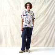 A person stands against a light-colored fabric backdrop, wearing the Far Afield Busey SS Shirt in Doodles Print, paired with dark pants and beige shoes.
