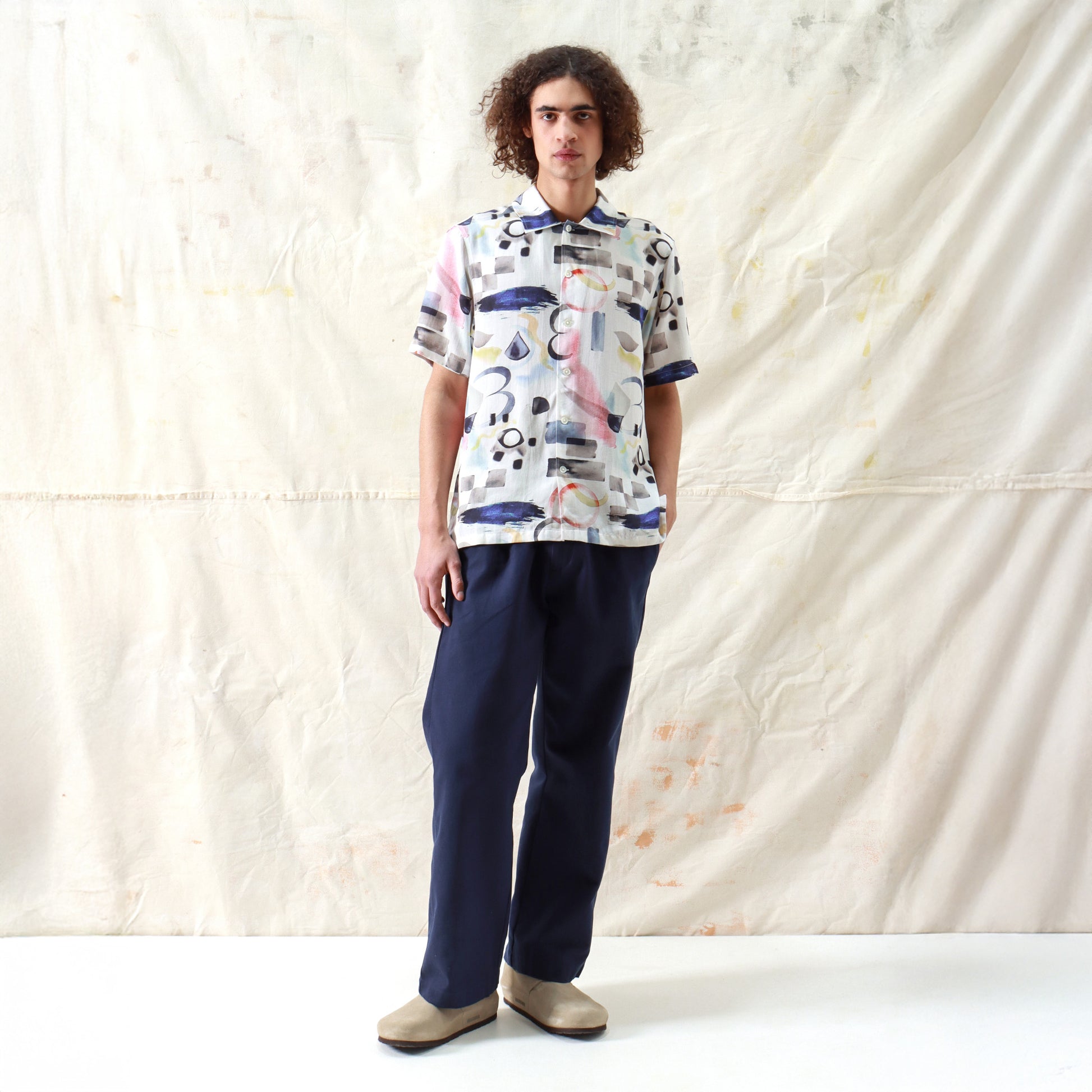 A person stands against a light-colored fabric backdrop, wearing the Far Afield Busey SS Shirt in Doodles Print, paired with dark pants and beige shoes.