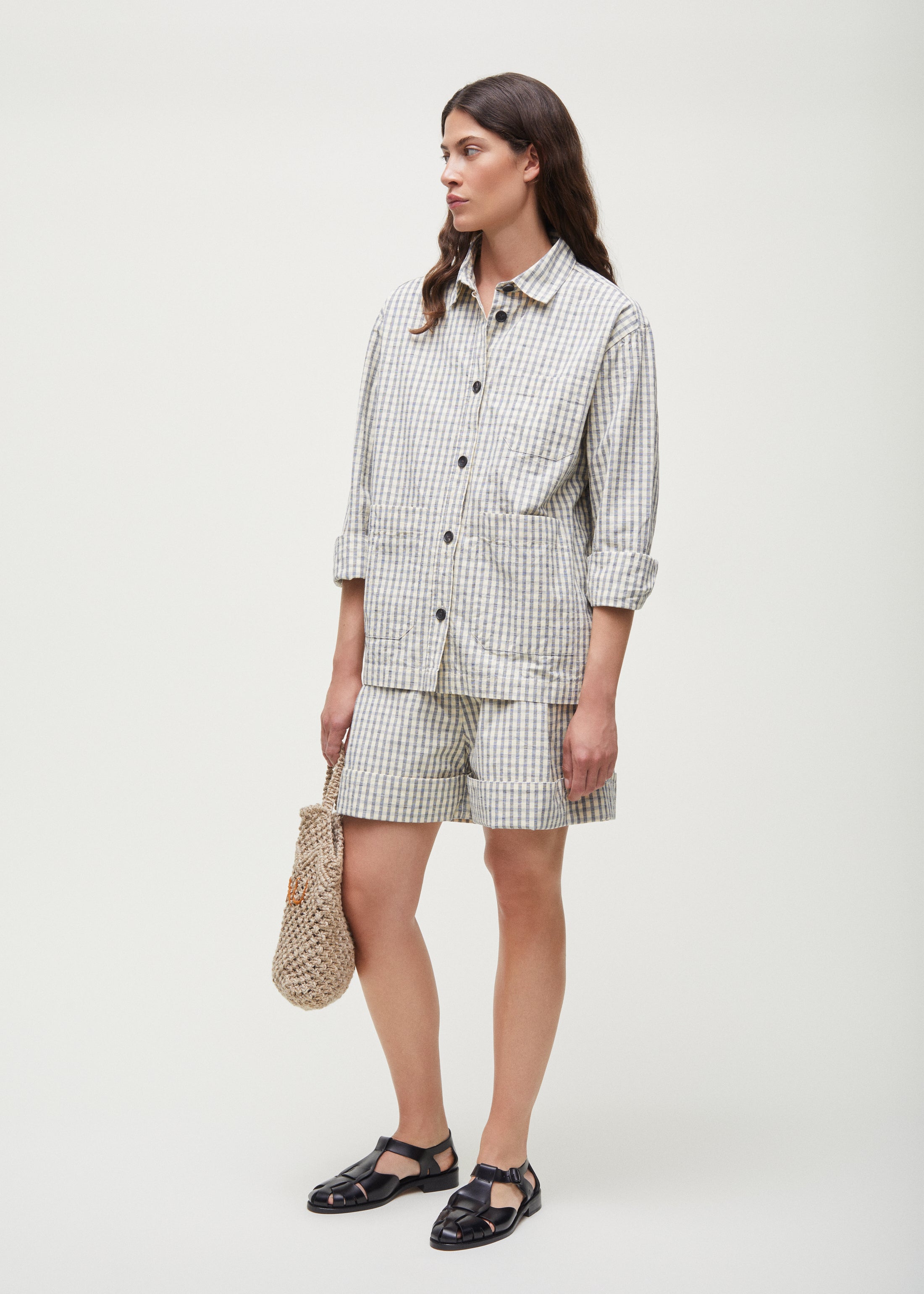 A person wearing the Aiayu Ashly Shirt Check and shorts set, holding a woven bag with handmade horn buttons, stands sideways against a plain background.