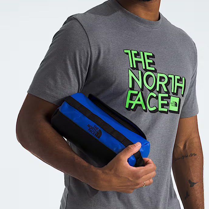 A person wearing a grey The North Face t-shirt holds a blue The North Face Base Camp Travel Canister S, made with recycled material, against their side.