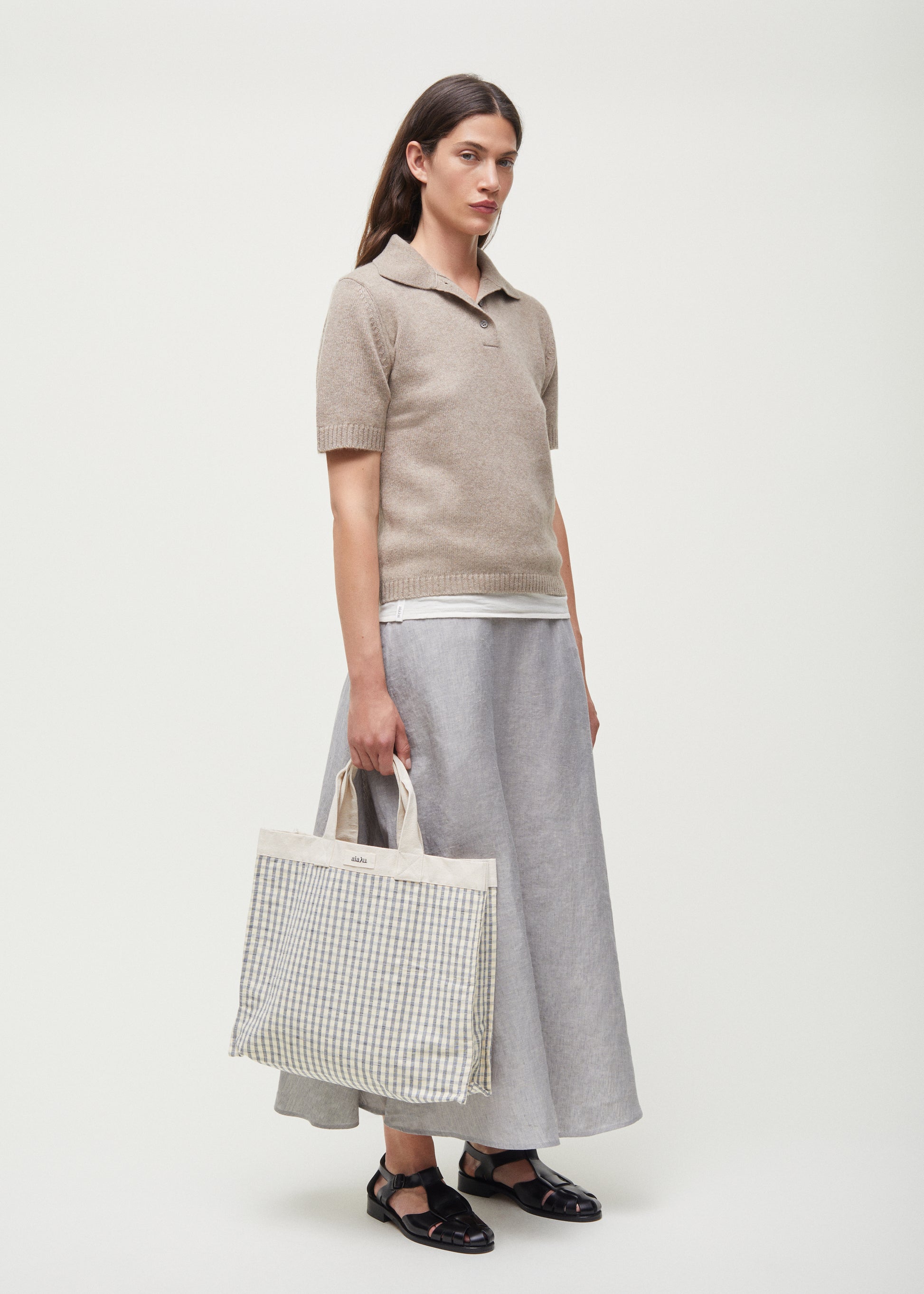 A person in a beige polo sweater, Aiayu Bea Skirt Linen in gray, and black sandals holds a checkered tote bag against a plain background.