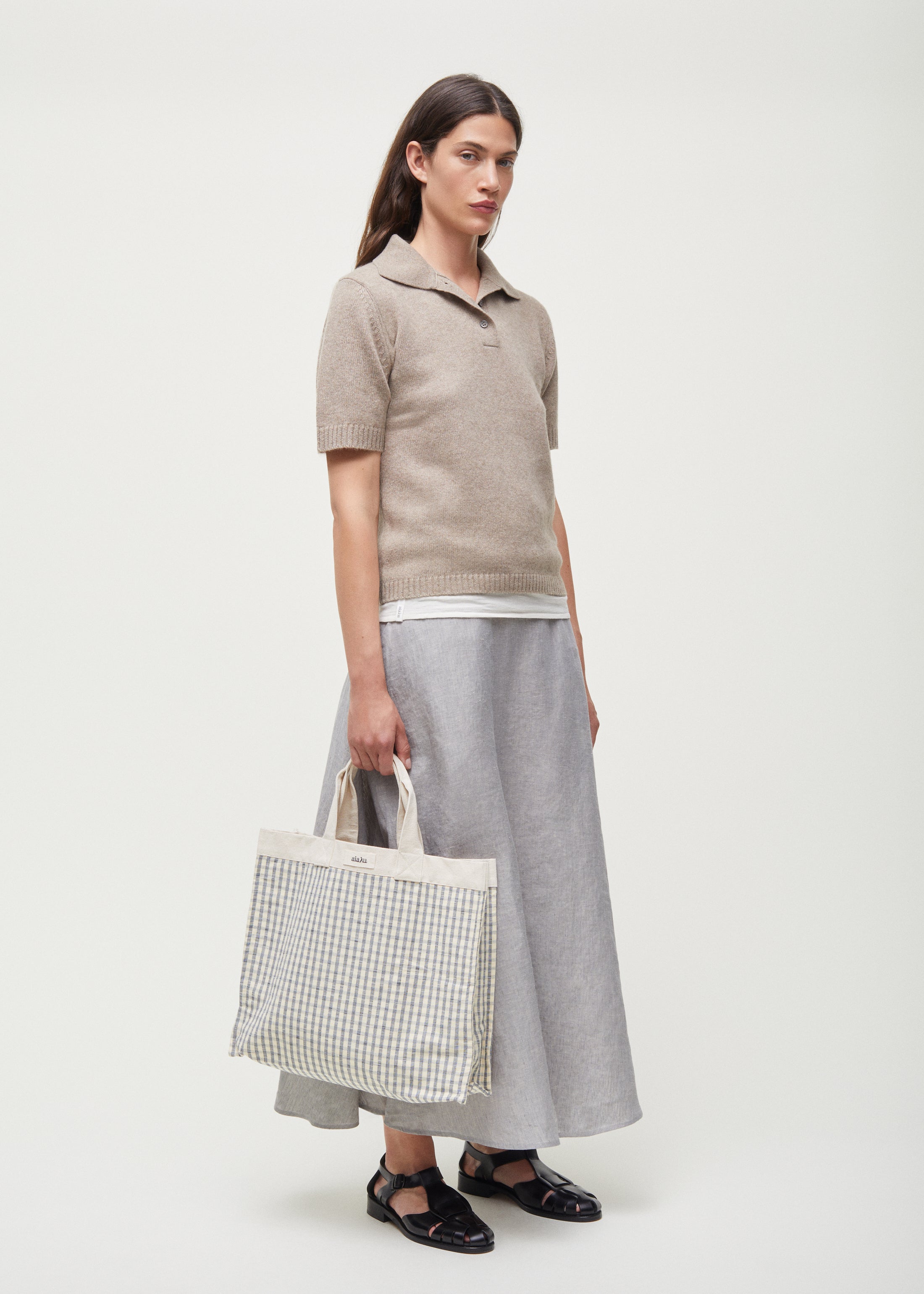 A person in a beige polo sweater, Aiayu Bea Skirt Linen in gray, and black sandals holds a checkered tote bag against a plain background.