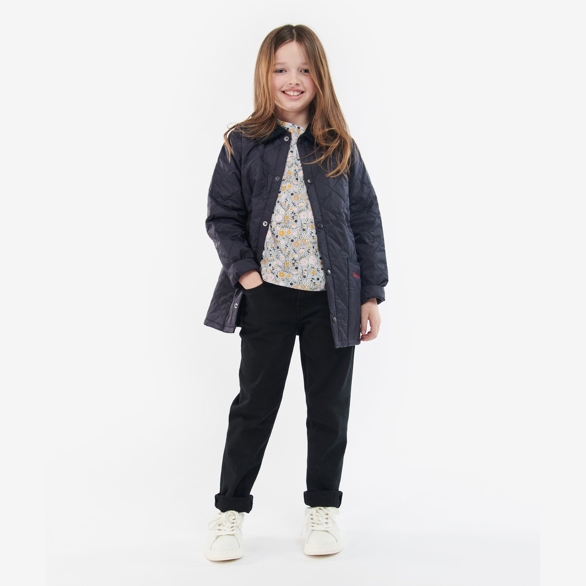 A young girl with long hair stands smiling against a white background, wearing the Barbour Boys Liddesdale Quilted Jacket with a needlecord collar, a floral shirt, black pants, and white sneakers.