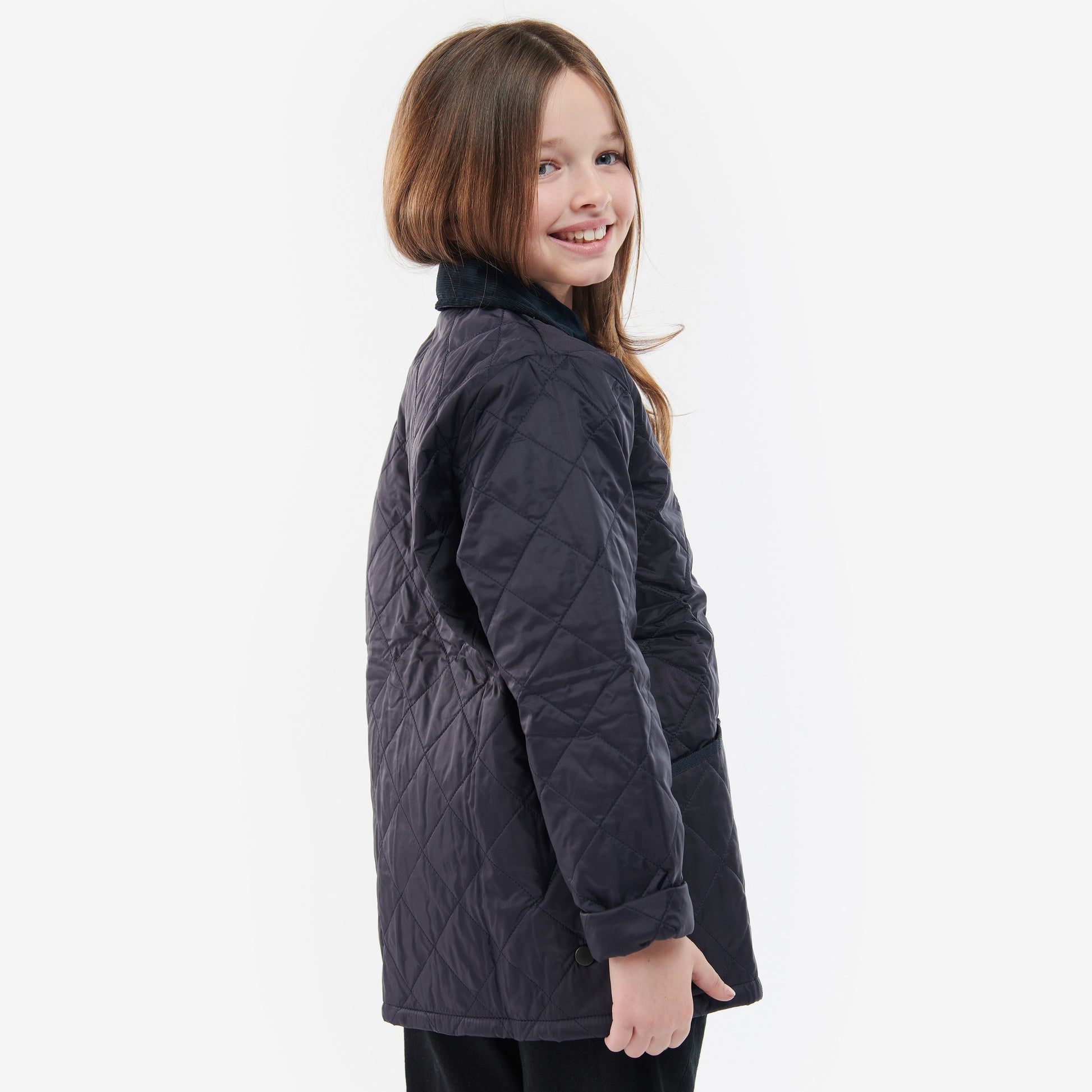 A girl with long hair smiles, wearing the Barbour Boys Liddesdale Quilted Jacket with a needlecord collar, turned slightly to her side against a plain background.