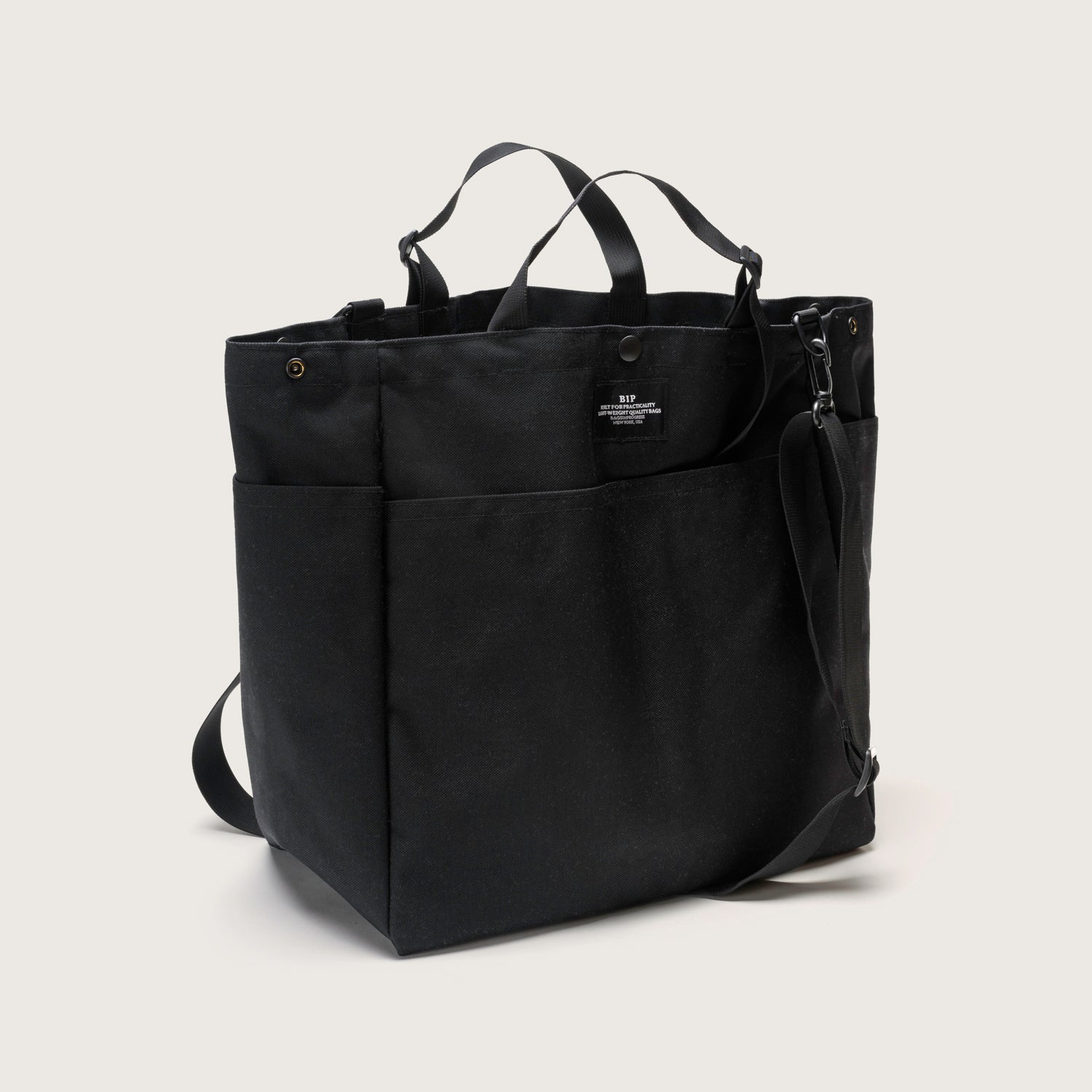 The Carry All Beach Bag by Bags in Progress is a black canvas tote with side pockets, adjustable handles, and a detachable shoulder strap, shown against a plain light background.