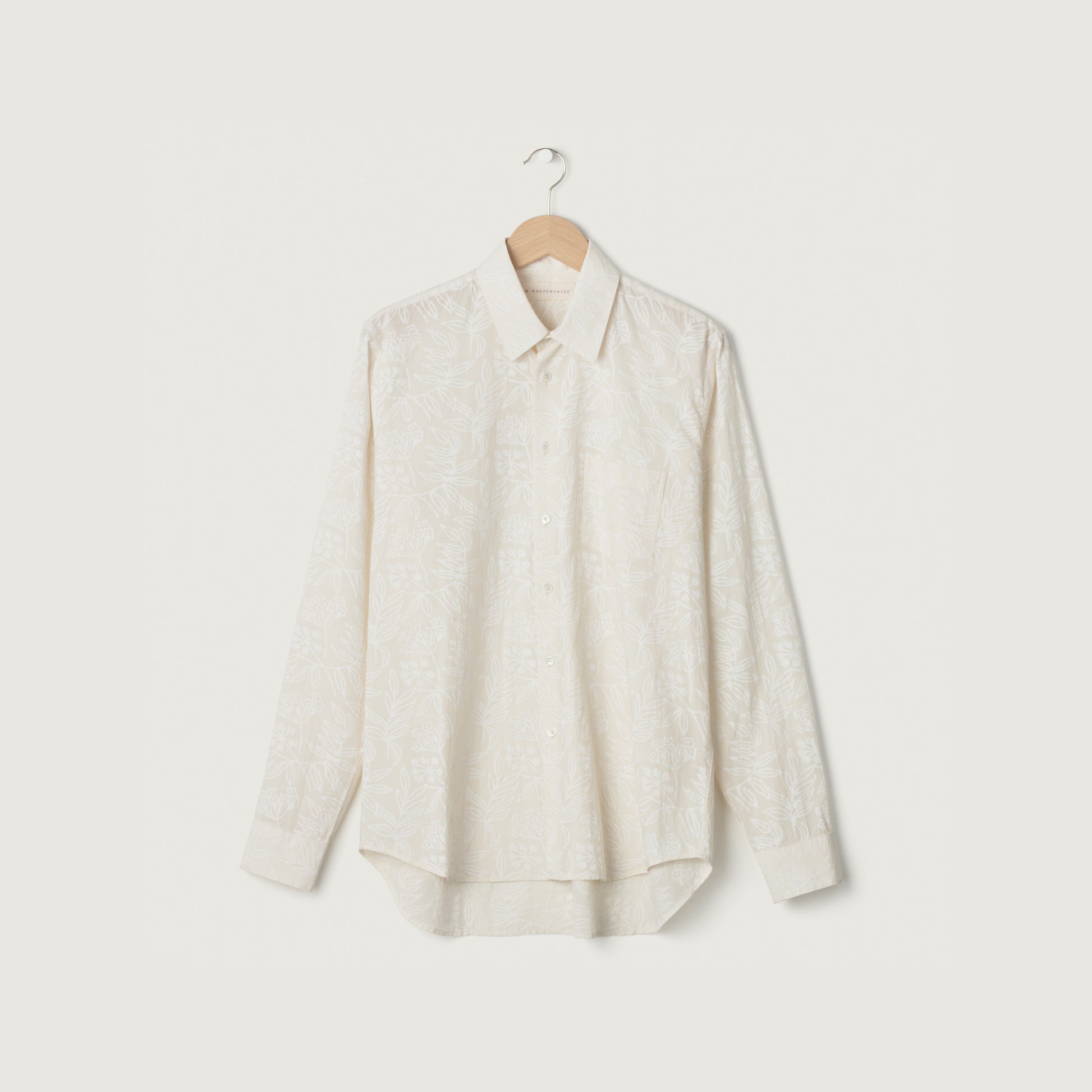 The Chris Shirt, Negative Floral by Jan Machenhauer—a long-sleeve, button-up shirt with a subtle white floral pattern—is shown on a wooden hanger against a plain white background.