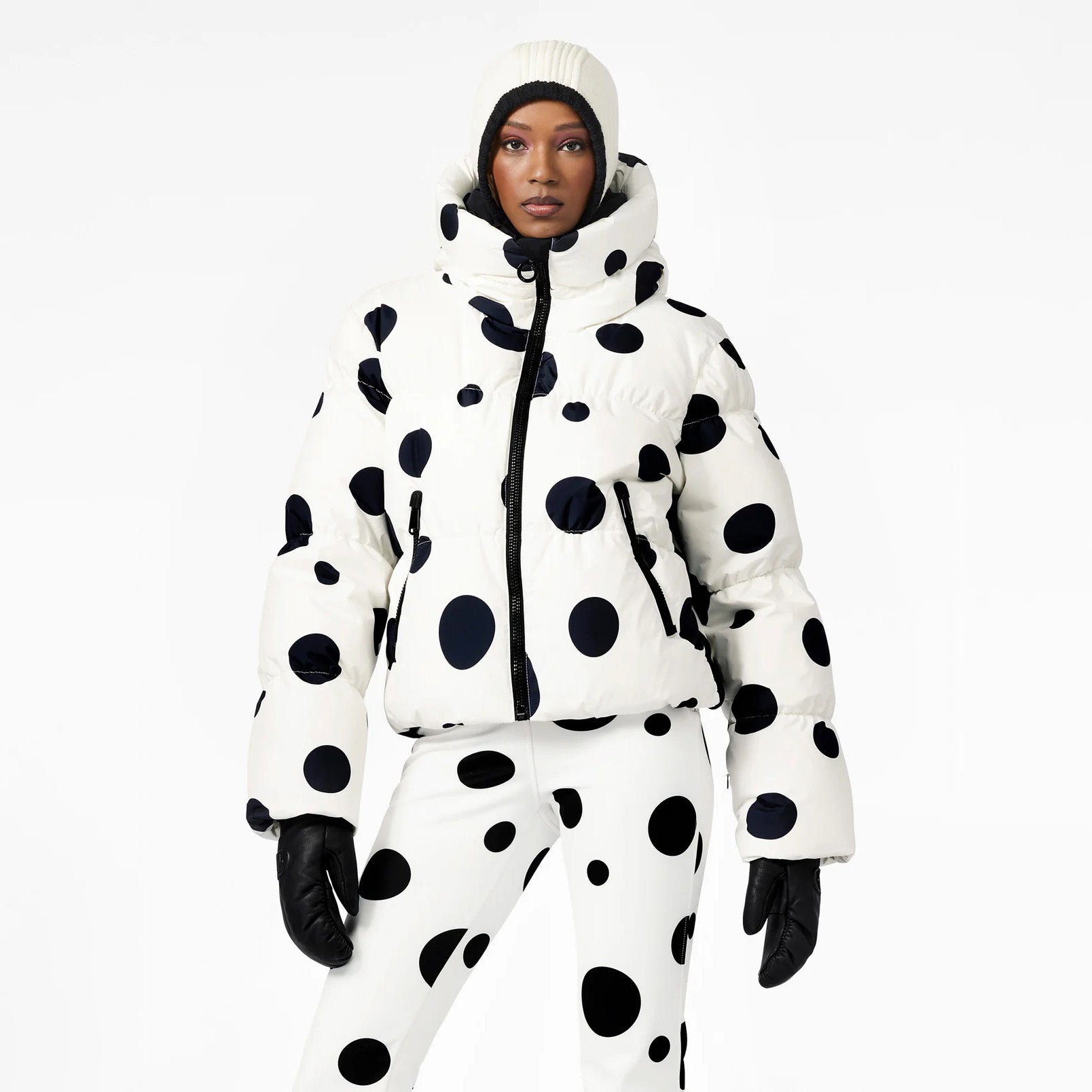 Against a plain backdrop, a person wears Goldbergh's Grand Ski Jacket in Dot Cream with matching pants. Completing the look with black gloves and a white beanie, it highlights insulated ski apparel made from premium waterproof fabric.