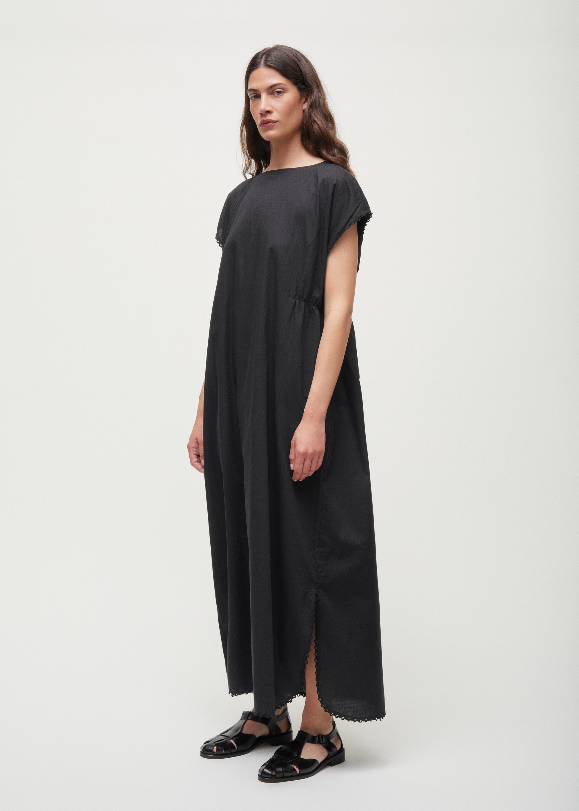Against a plain backdrop, a person dons the Aiayu Diamond Dress, a sleeveless, loose-fitting maxi crafted from GOTS-certified cotton, paired with black sandals.