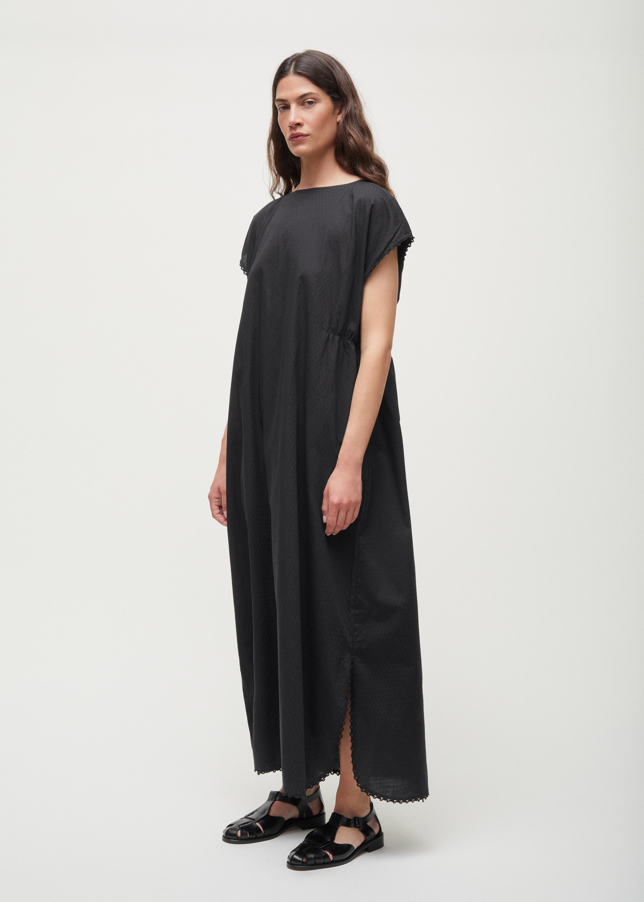 Against a plain backdrop, a person dons the Aiayu Diamond Dress, a sleeveless, loose-fitting maxi crafted from GOTS-certified cotton, paired with black sandals.