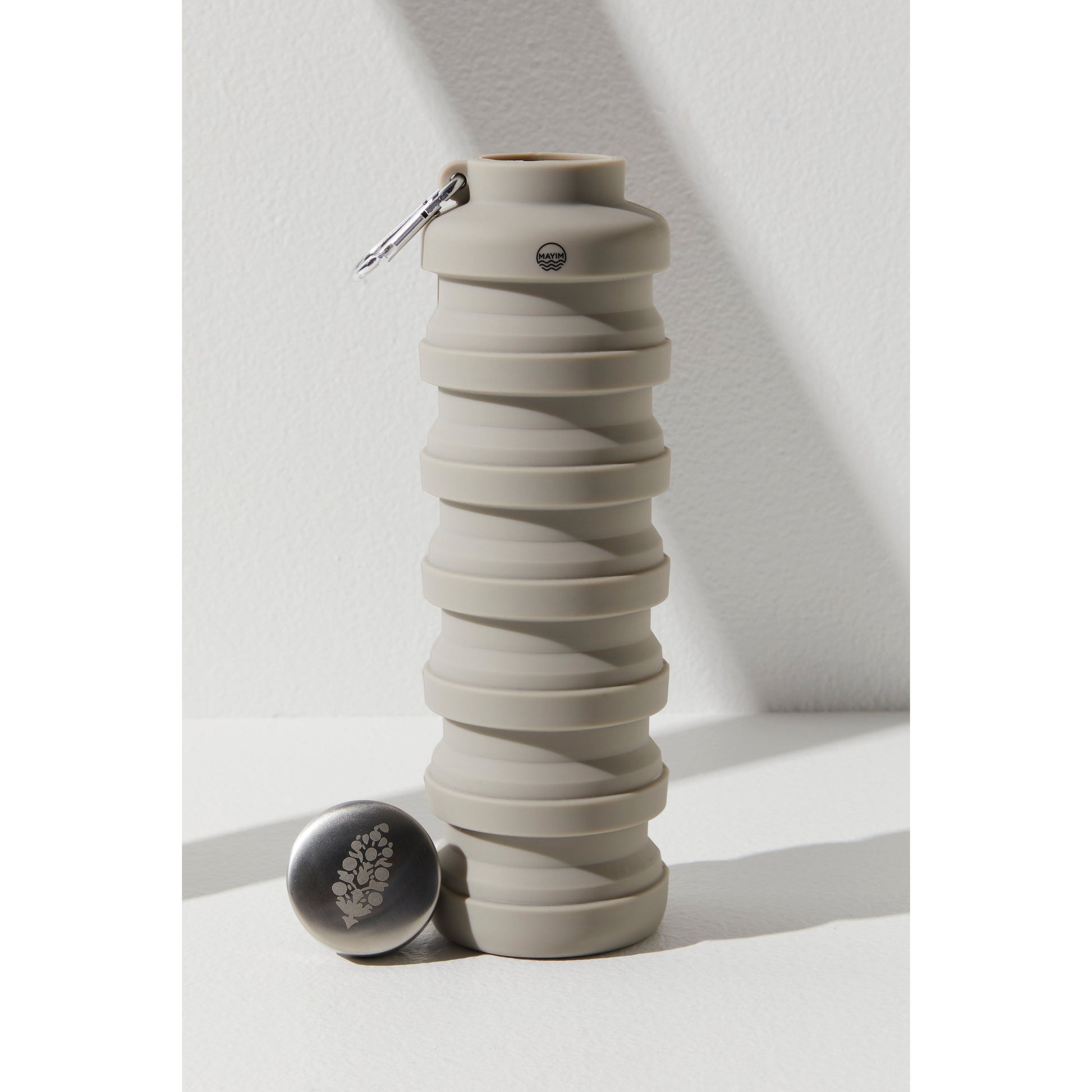 A collapsible, grey silicone Free People Movement 24OZ Carabiner Bottle with its lid off, displayed on a white surface under natural light, casting a soft shadow.