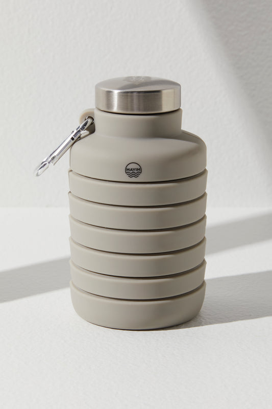 A collapsible, gray silicone 24OZ Carabiner Bottle by Free People Movement with a stainless steel cap and a carabiner attached, displayed against a white textured background.