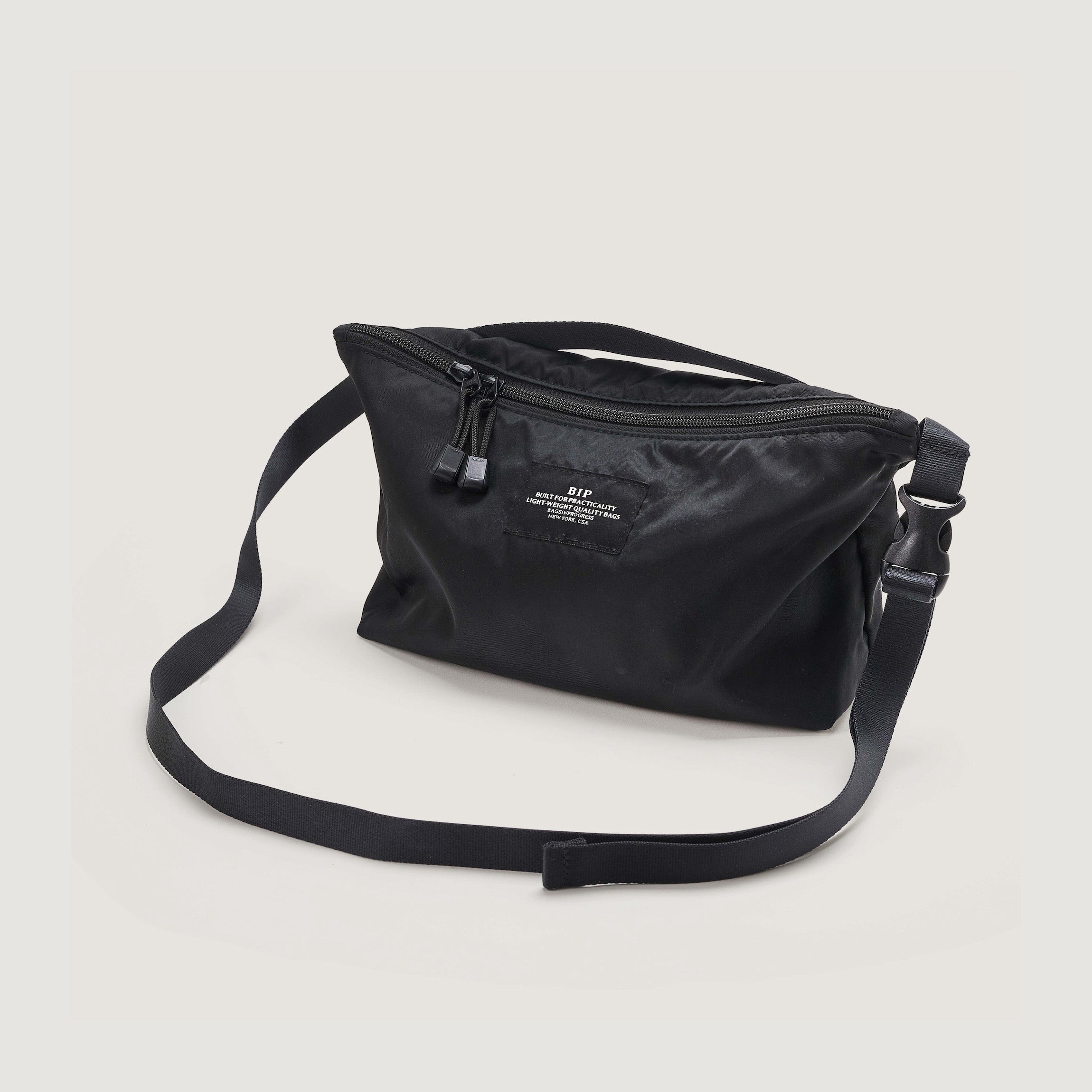 The Bags in Progress Fannypack Crossbody is a black recycled polyester bag with a zippered top, adjustable strap, and small front label—shown against a plain light background.