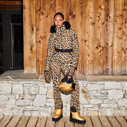 A person stands against a wooden wall wearing Goldbergh's breathable Minou Ski Pants in a leopard print (Jaguar), accessorized with a black belt, gloves, and gold boots while holding a matching gold helmet with black details.