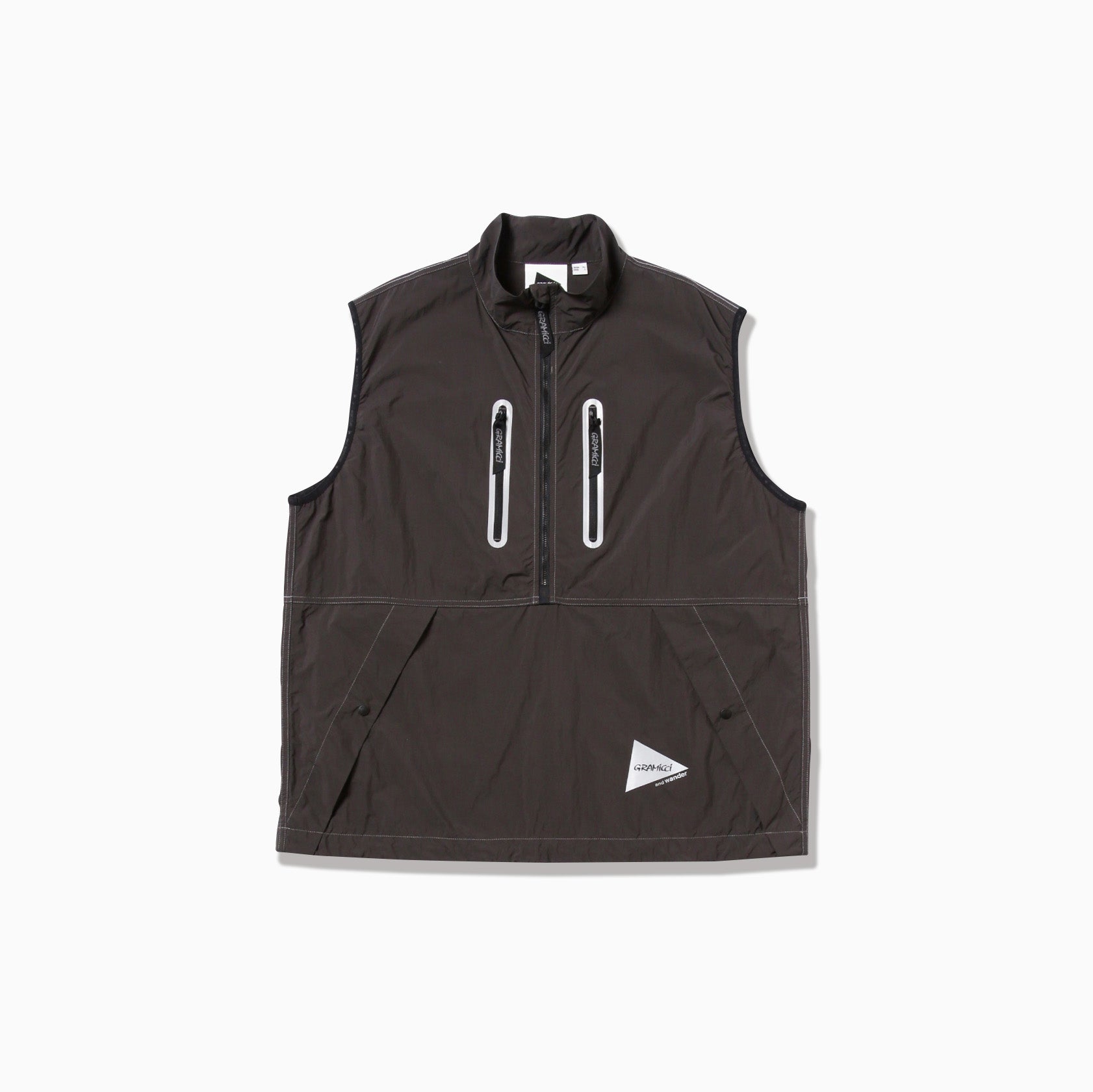 The Brushed Nylon Vest by Gramicci x And Wander is a dark gray zip-up with a high collar, two vertical chest pockets, and a triangular logo patch at the bottom.