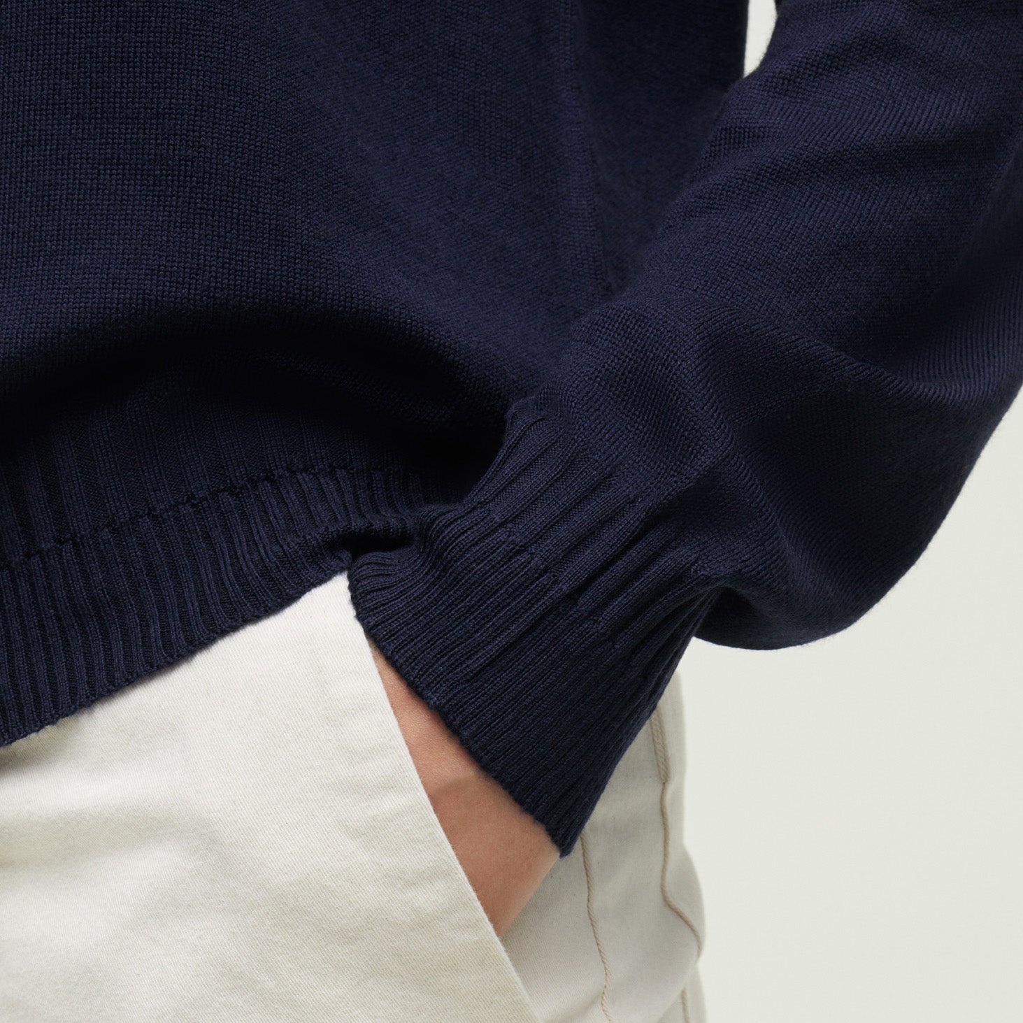 A person wearing the Aiayu Gina Sweater in navy blue, made of 100% wool, casually tucks one hand into the pocket of cream-colored pants.