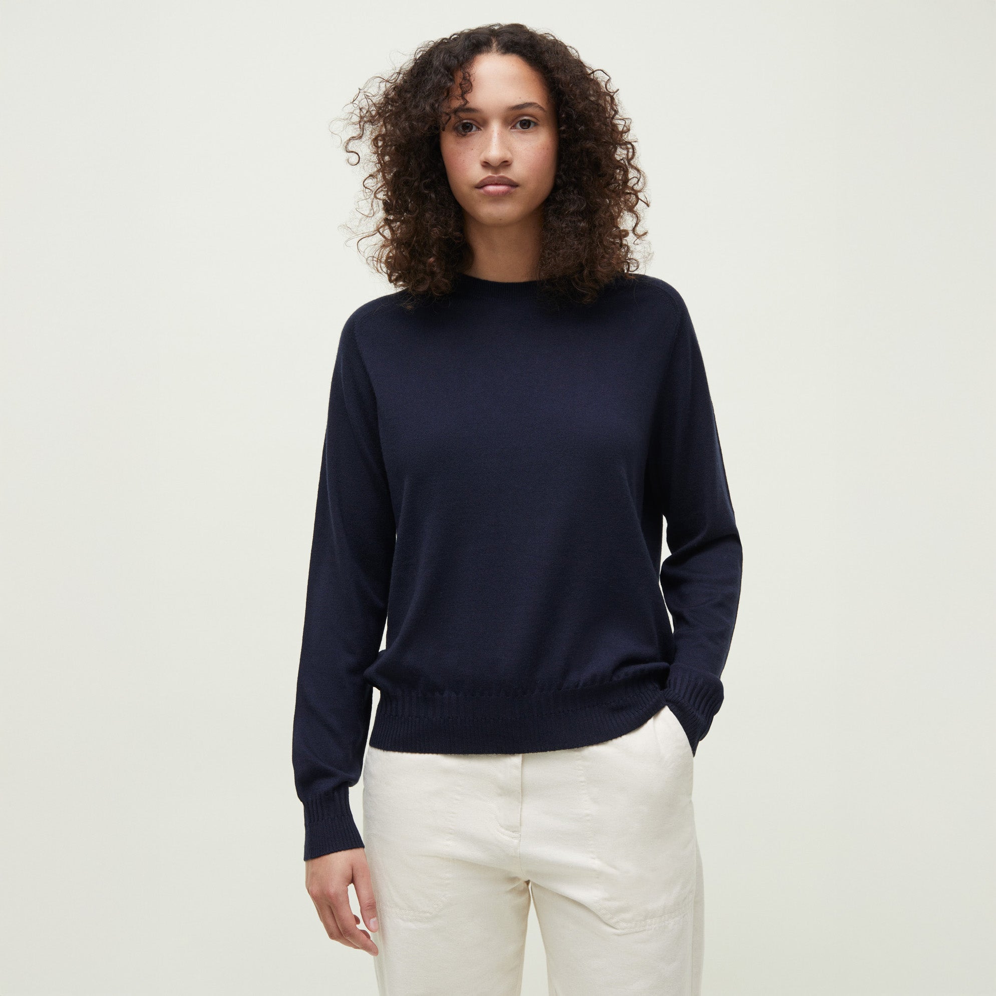 A person with curly hair is wearing the Gina Sweater by Aiayu, a navy merino wool piece, paired with cream pants while standing against a plain light background.