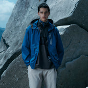 Someone in a Gramicci x And Wander Brushed Nylon Jacket and light pants stands outdoors in front of large rocks.