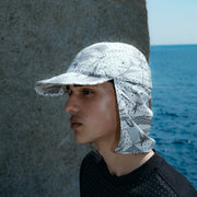 A person wearing a Gramicci x And Wander Brushed Nylon Sunshade Cap and a black shirt stands near a stone wall with the ocean in the background.