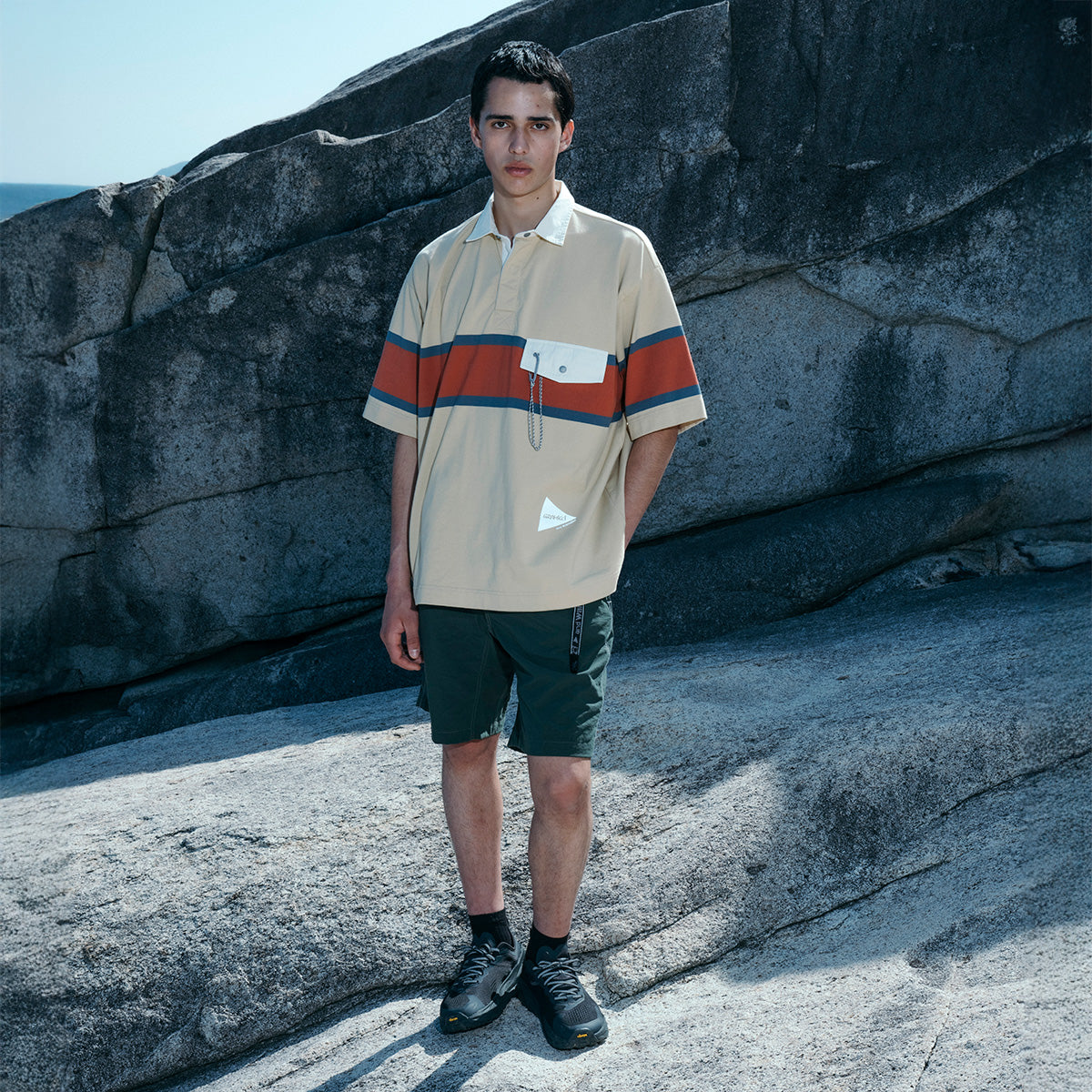 Person standing on rocky terrain wearing a Gramicci x And Wander Tech Rugby Shirt, green shorts, and black shoes.